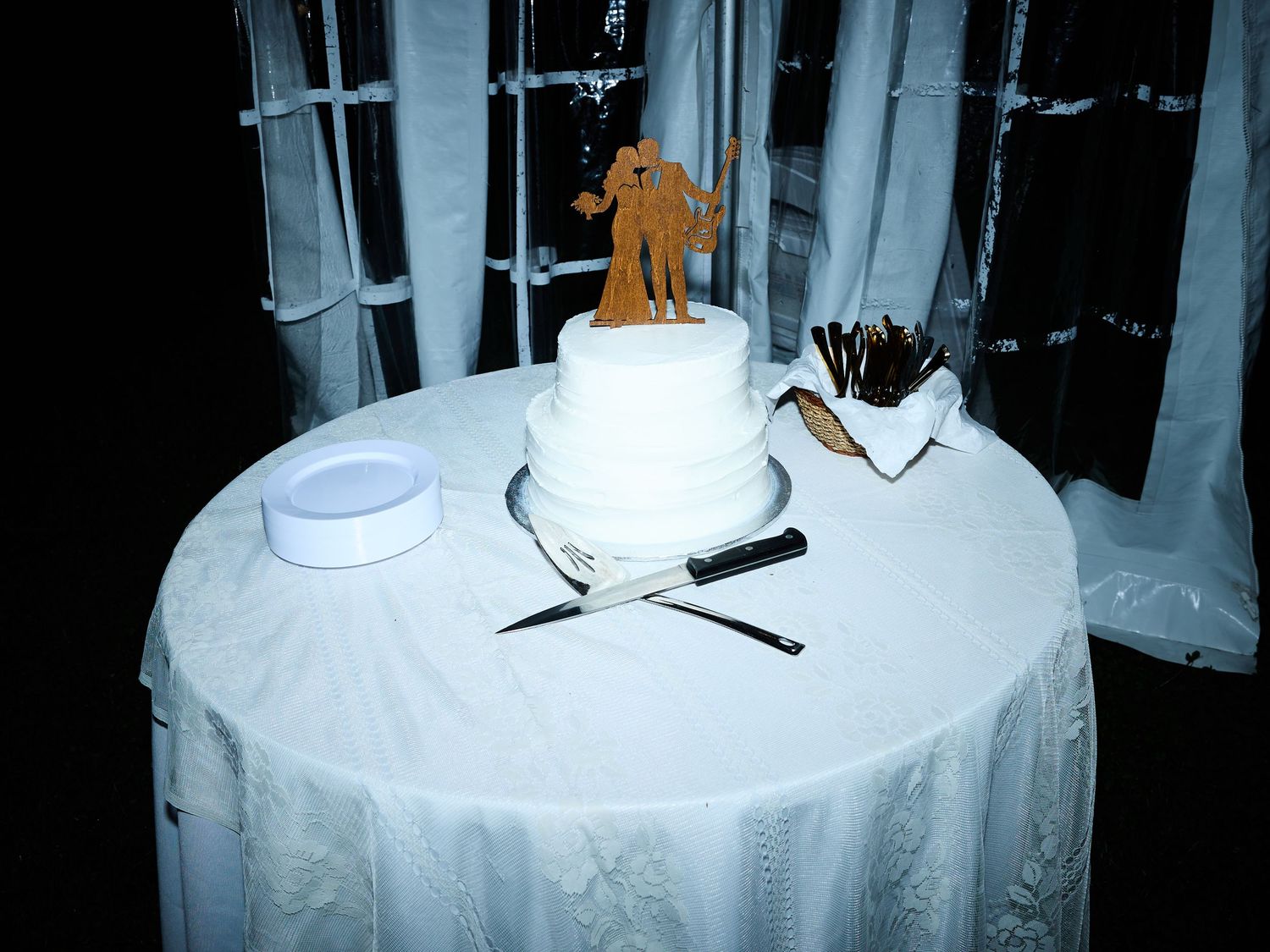 A white two-tier wedding cake with a wooden couple topper sits on a draped round table beside a cake knife and plates at night.