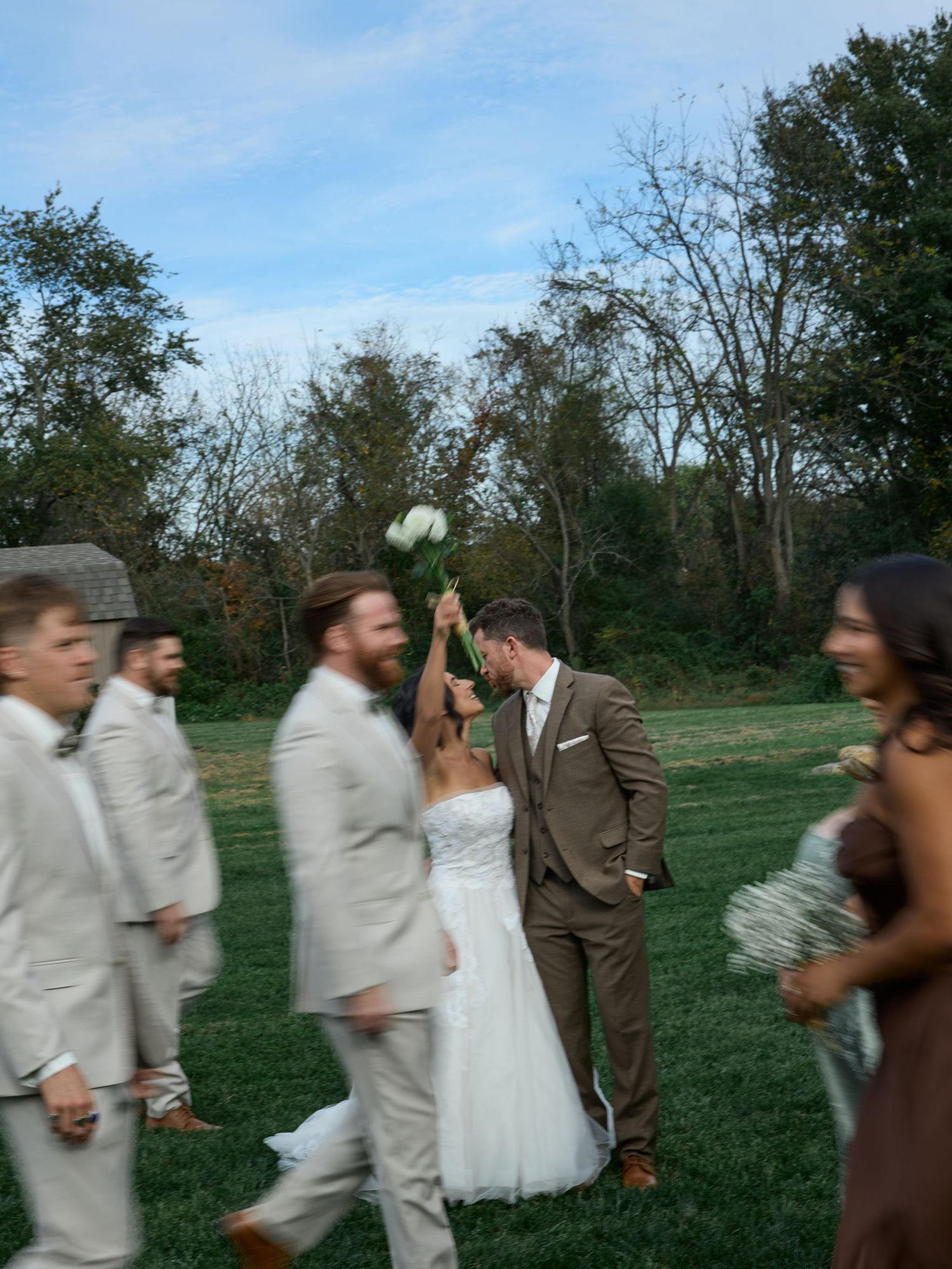 A bride and groom share a kiss on a grassy lawn while blurred groomsmen and a bridesmaid walk past in the foreground.