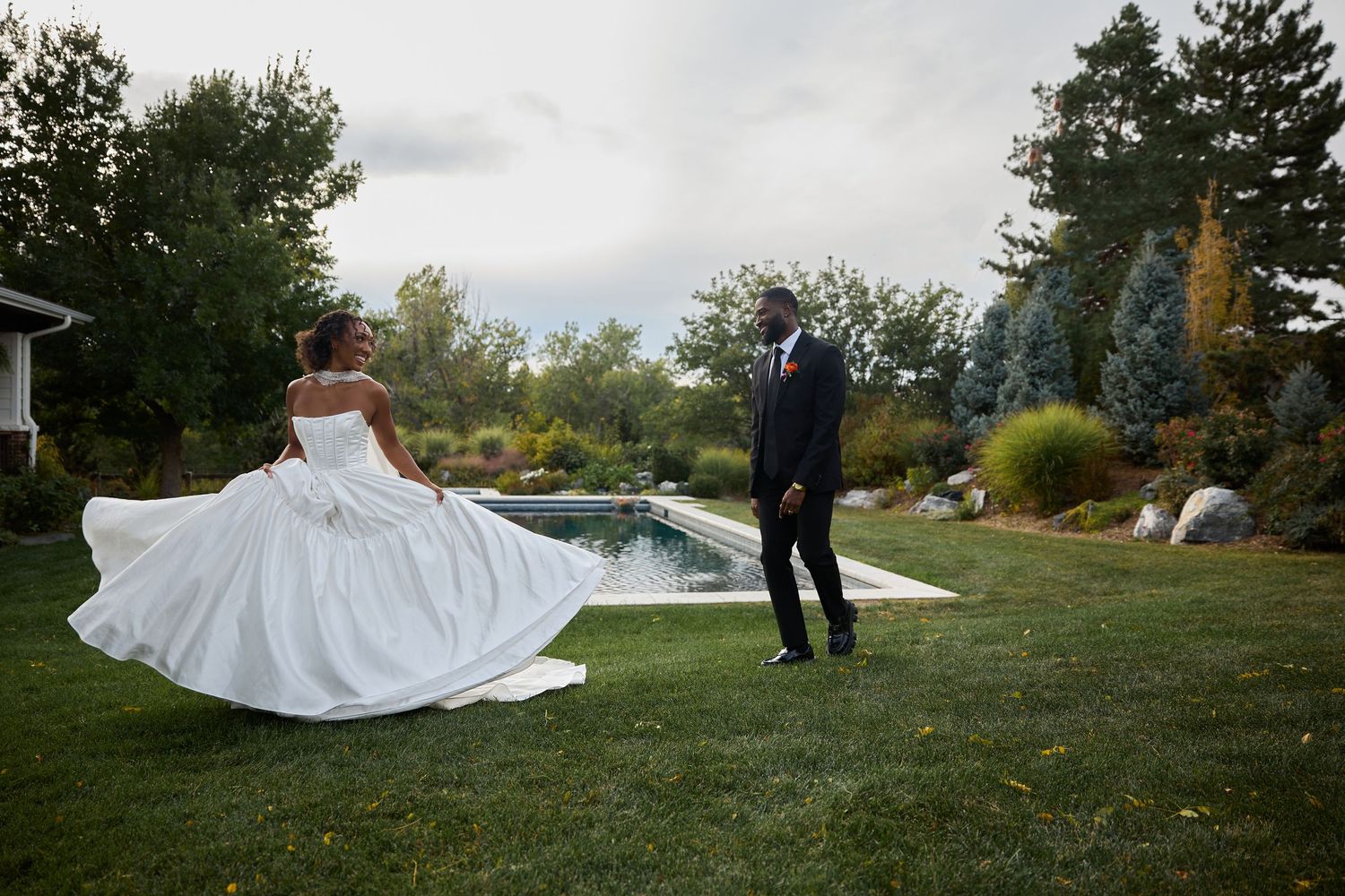 Bride spins in her full white wedding dress as her smiling groom in a dark suit looks on in a lush garden setting.