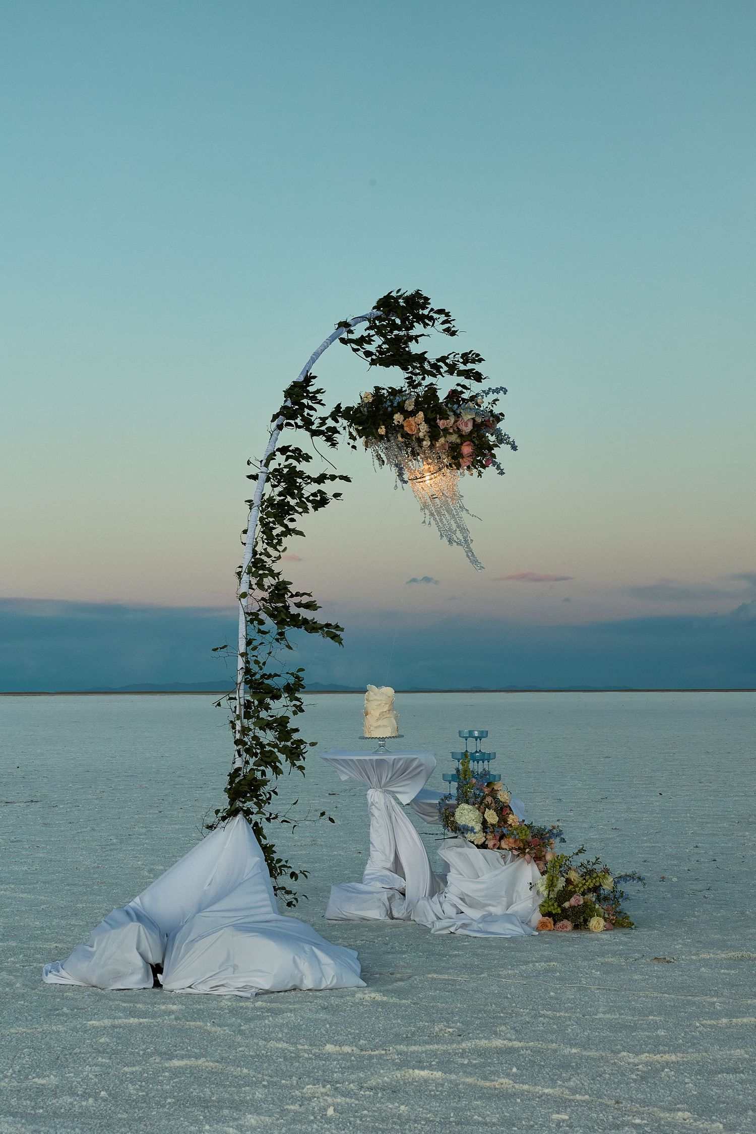 Romantic salt flat wedding setup with a floral arch, chandelier, cake table, and white pillows at dusk.