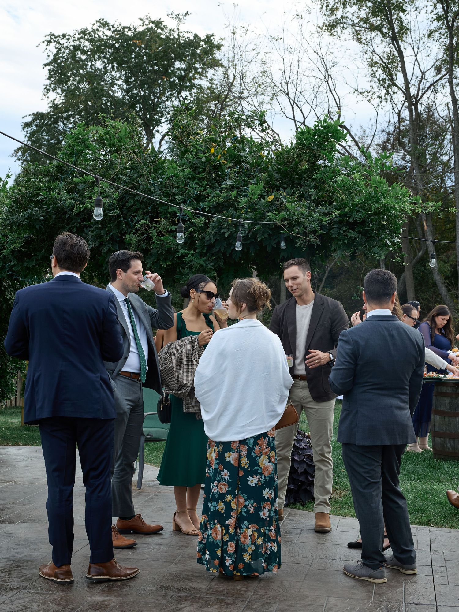 Wedding guests in formal attire mingle and chat on a stone patio beneath string lights and green trees at an outdoor reception.