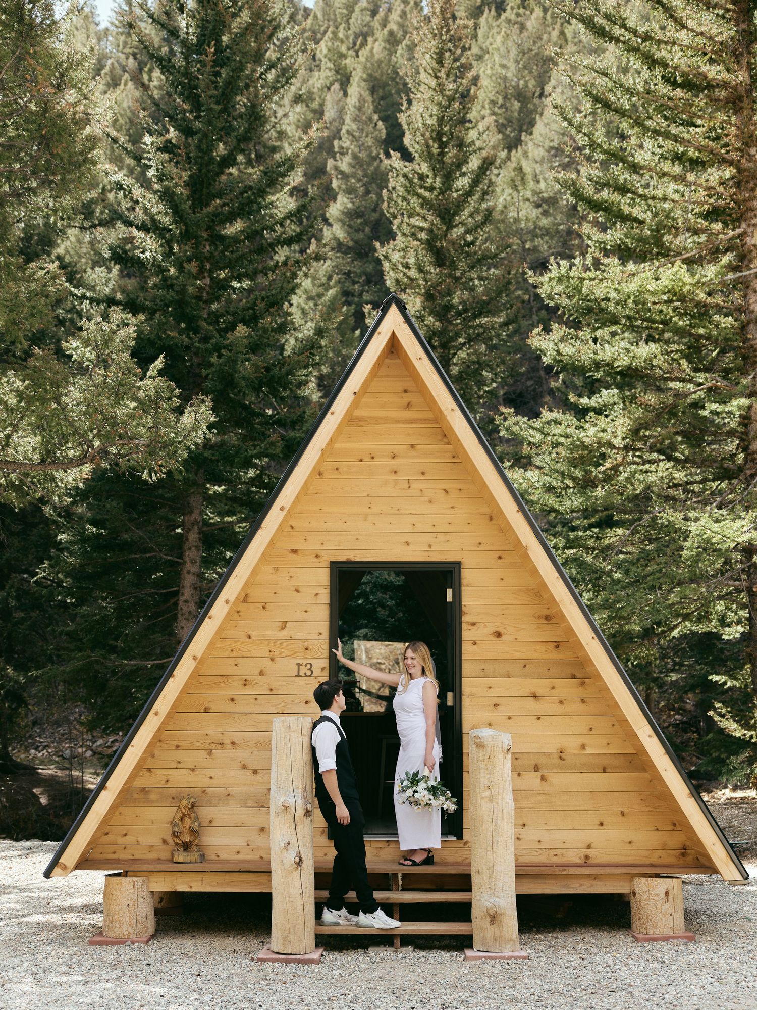 Bride and groom stand at the doorway of a wooden A-frame cabin surrounded by tall evergreen trees during a mountain elopement.