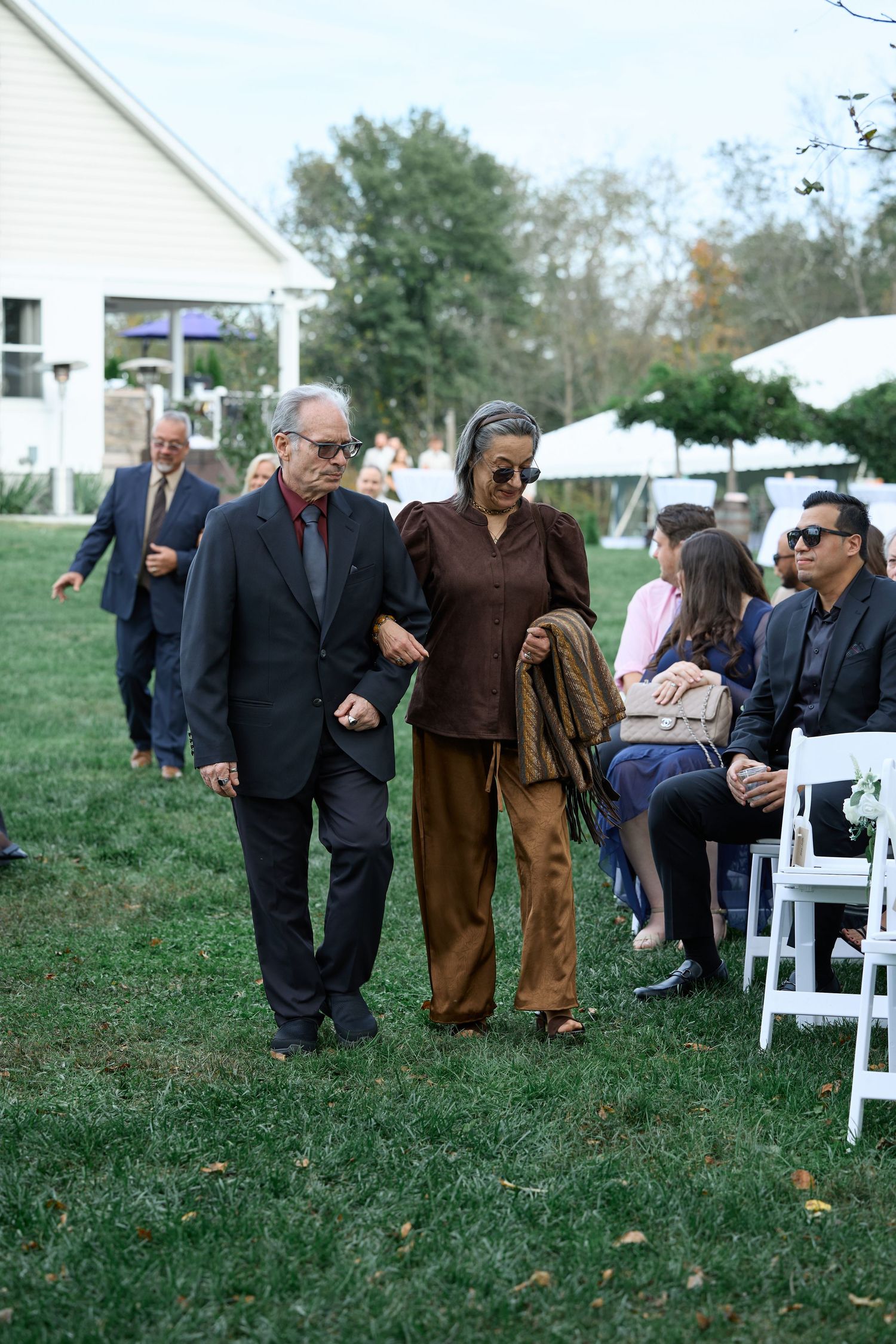 Older man in a dark suit escorts a woman in a brown outfit down the grass aisle at an outdoor wedding ceremony.