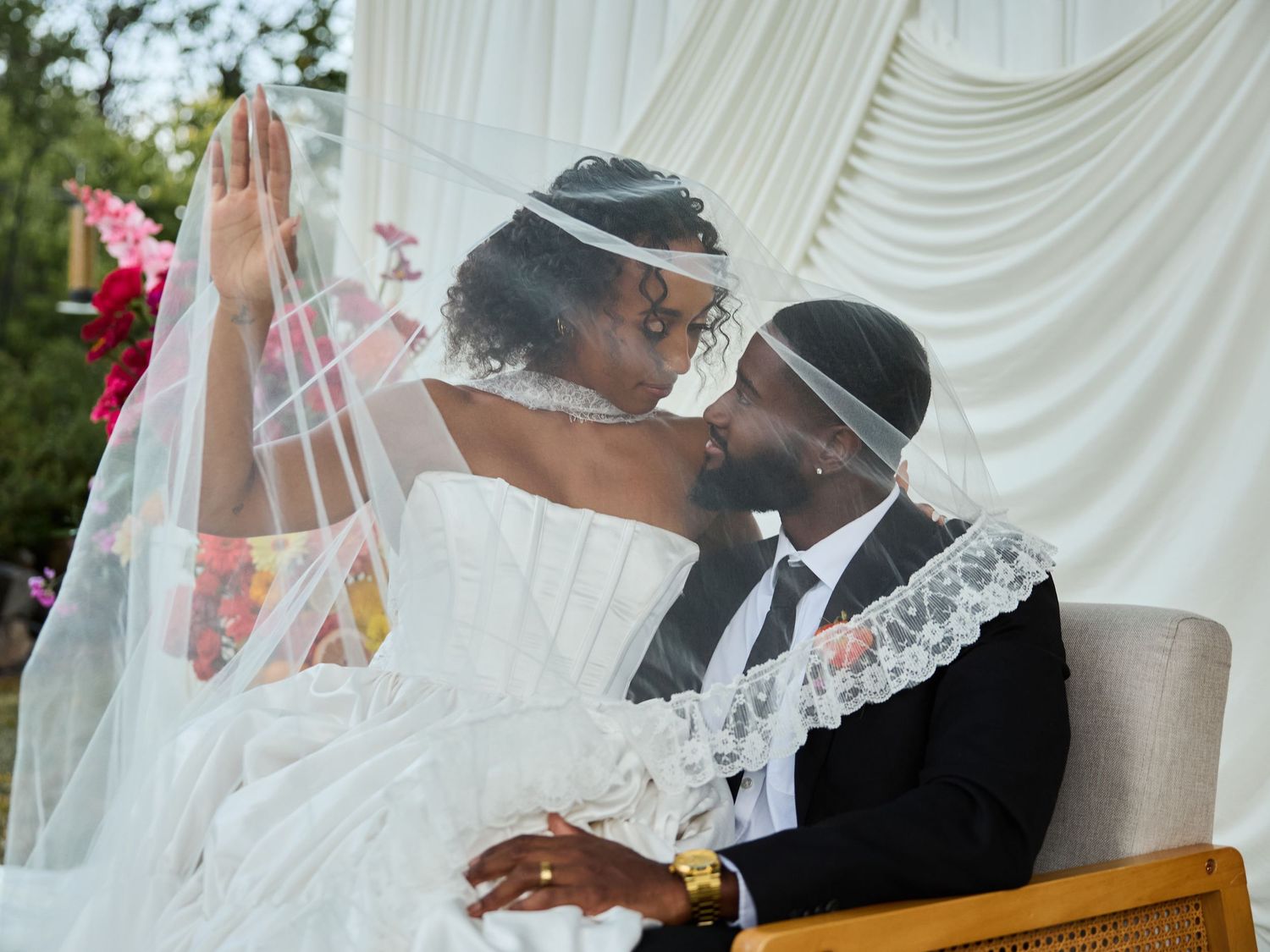 A bride and groom lean close under a lace-edged veil as she raises her hand, seated outdoors with white draping and flowers.