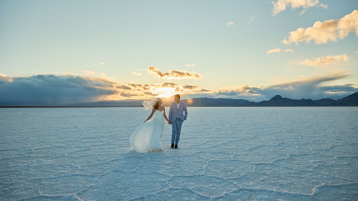Wedding couple holds hands on a glistening salt flat as the sun sets on the horizon, casting soft golden light across the sky.