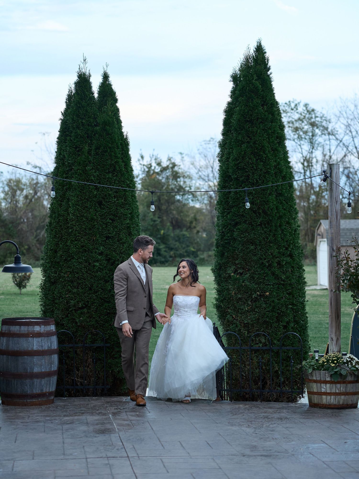 Bride in a strapless white gown and groom in a tan suit walk hand in hand between tall evergreens at an outdoor wedding venue.