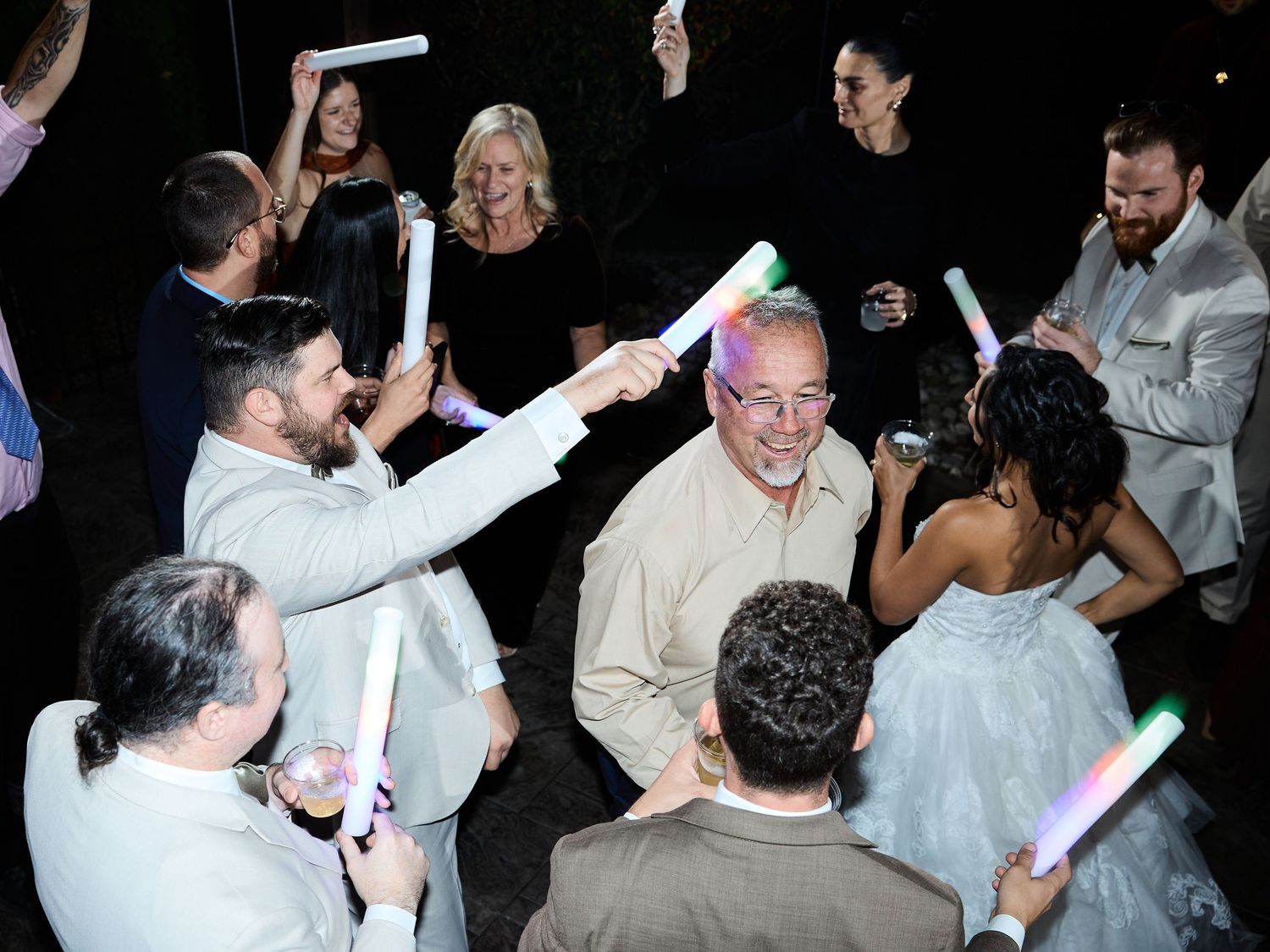 Wedding guests in suits and a bride in a white gown dance together under glowing foam light sticks at an evening reception.