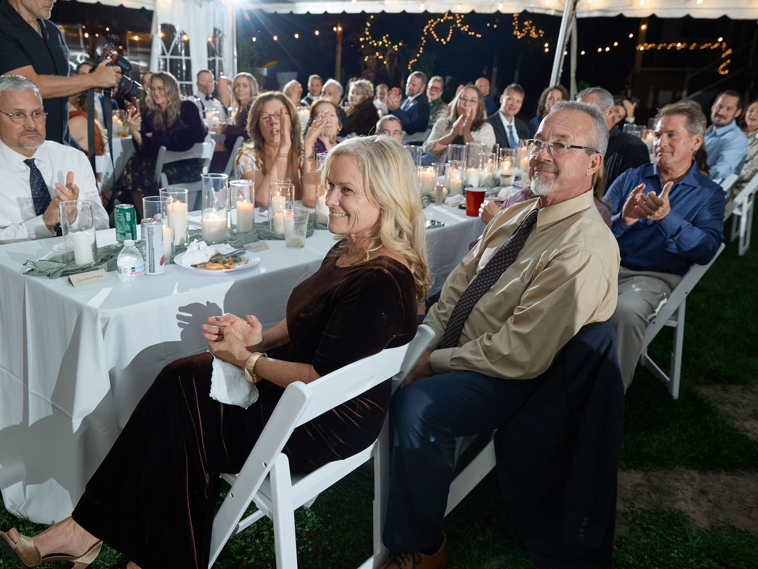 A smiling blonde woman and a man in glasses sit in white chairs at a wedding reception while other guests look on behind them.