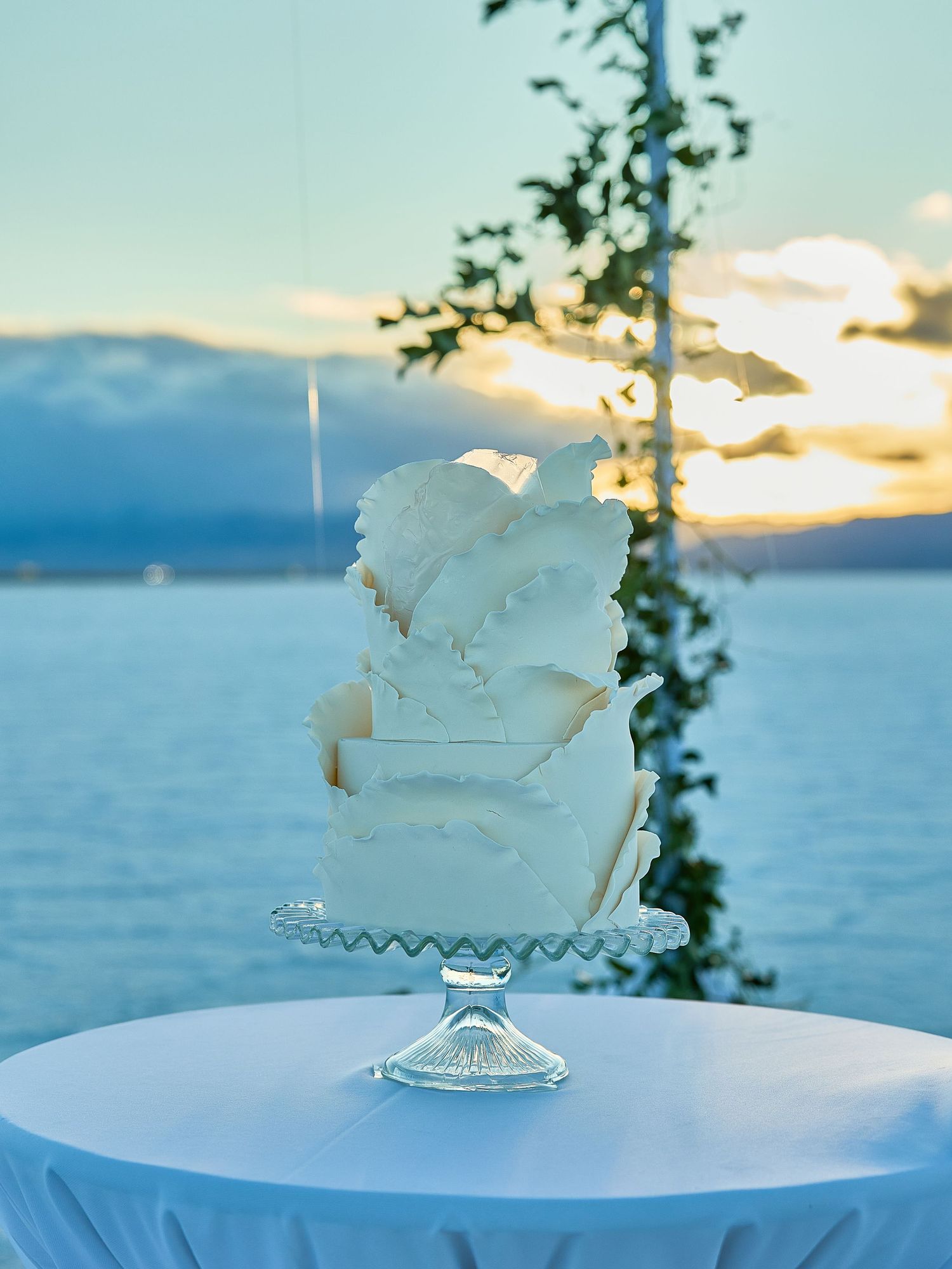 Sculptural white wedding cake with ruffled petal details on a glass stand, with a glowing sunset salt flat behind it.