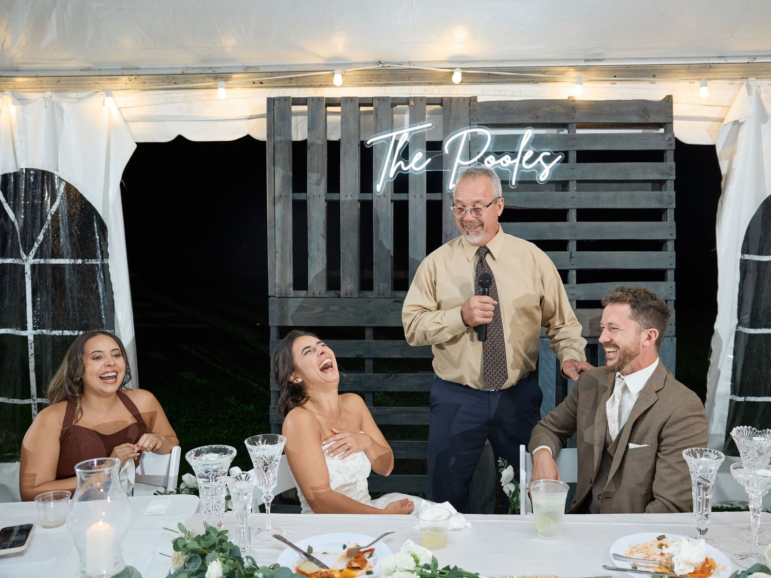 The bride laughs with her head tilted back as a man in glasses gives a toast beside the smiling groom at the wedding reception.