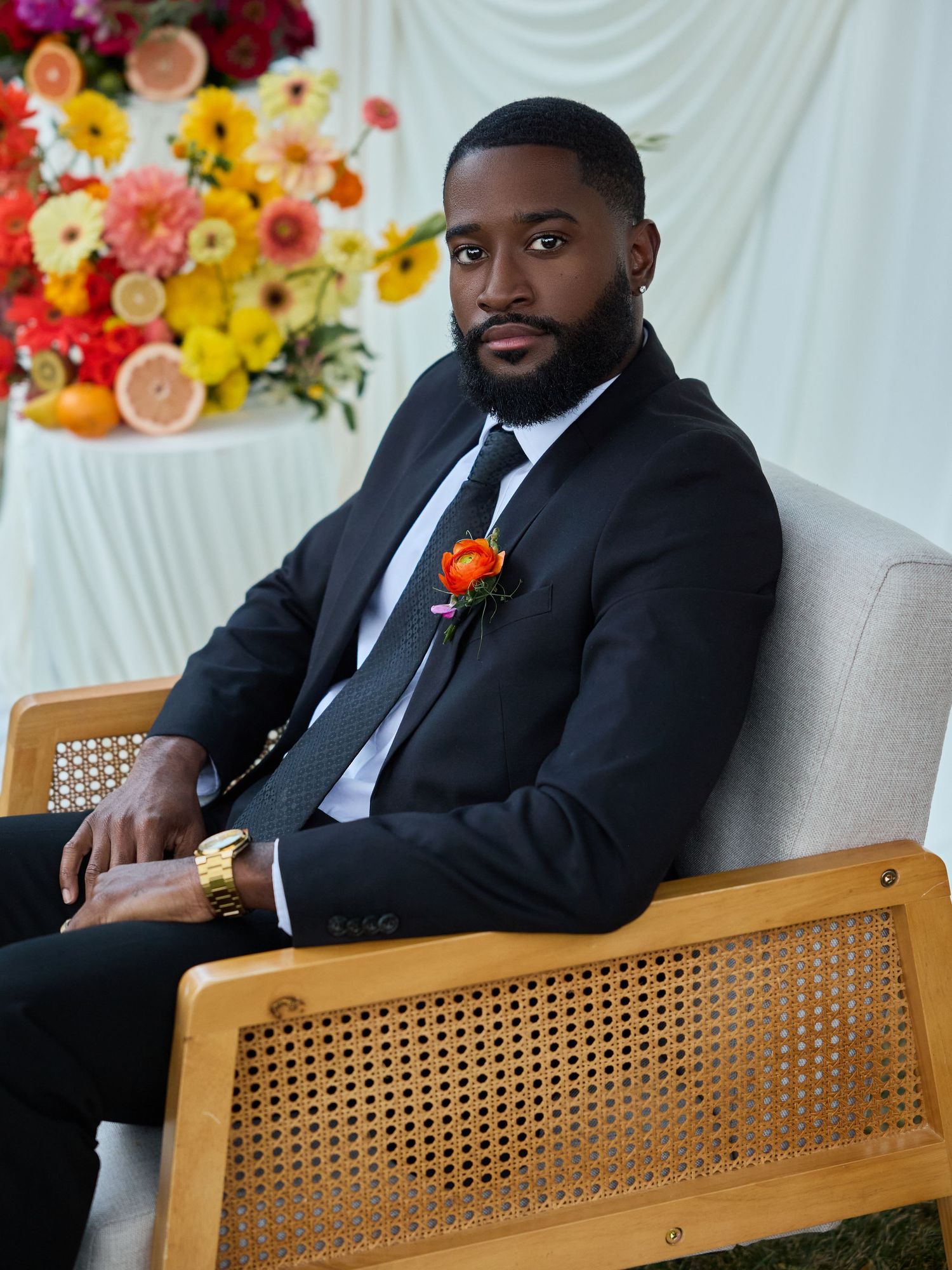 A groom in a black suit with an orange boutonniere sits in a wooden cane chair beside a table of citrus and flowers.