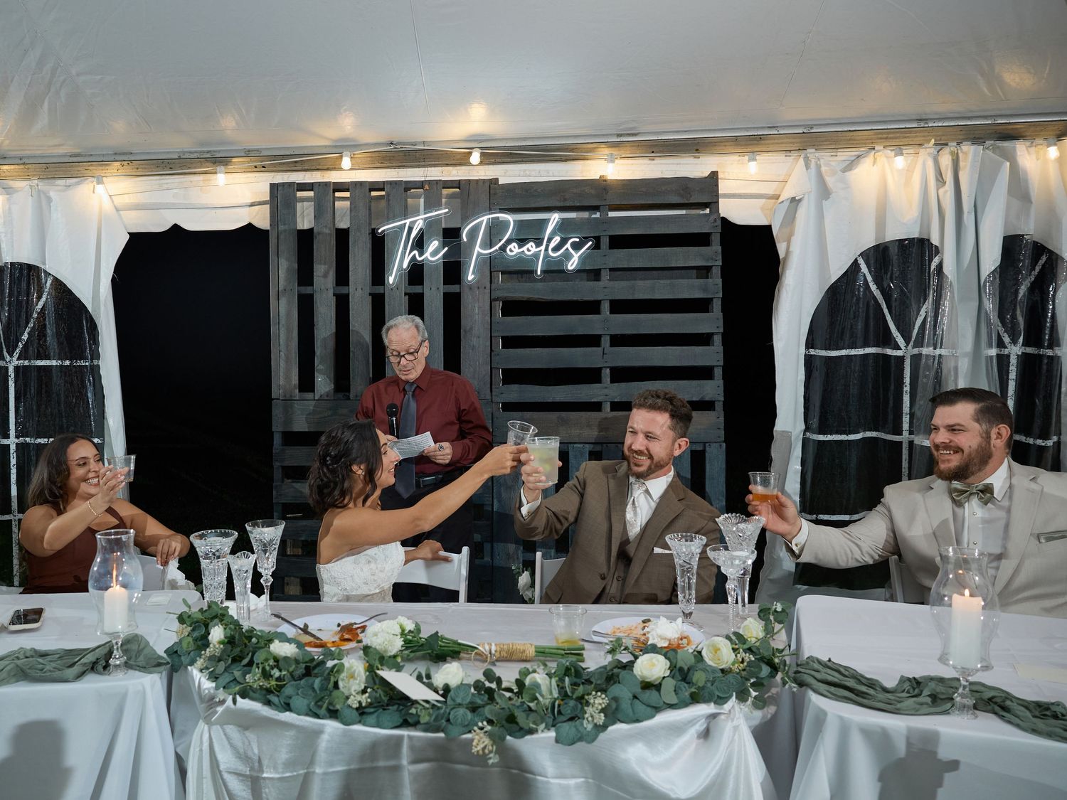 Wedding guests raise glasses in a toast at the head table beneath a tent, with a neon The Pooles sign behind the couple.