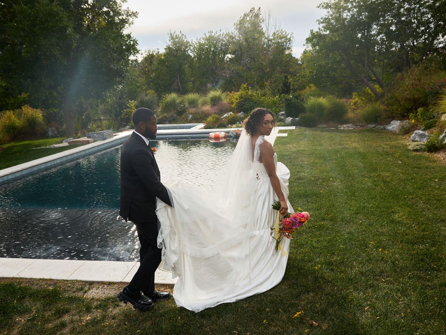 A groom in a dark suit walks beside his bride, whose long white veil and gown trail across the lawn near a garden pool.