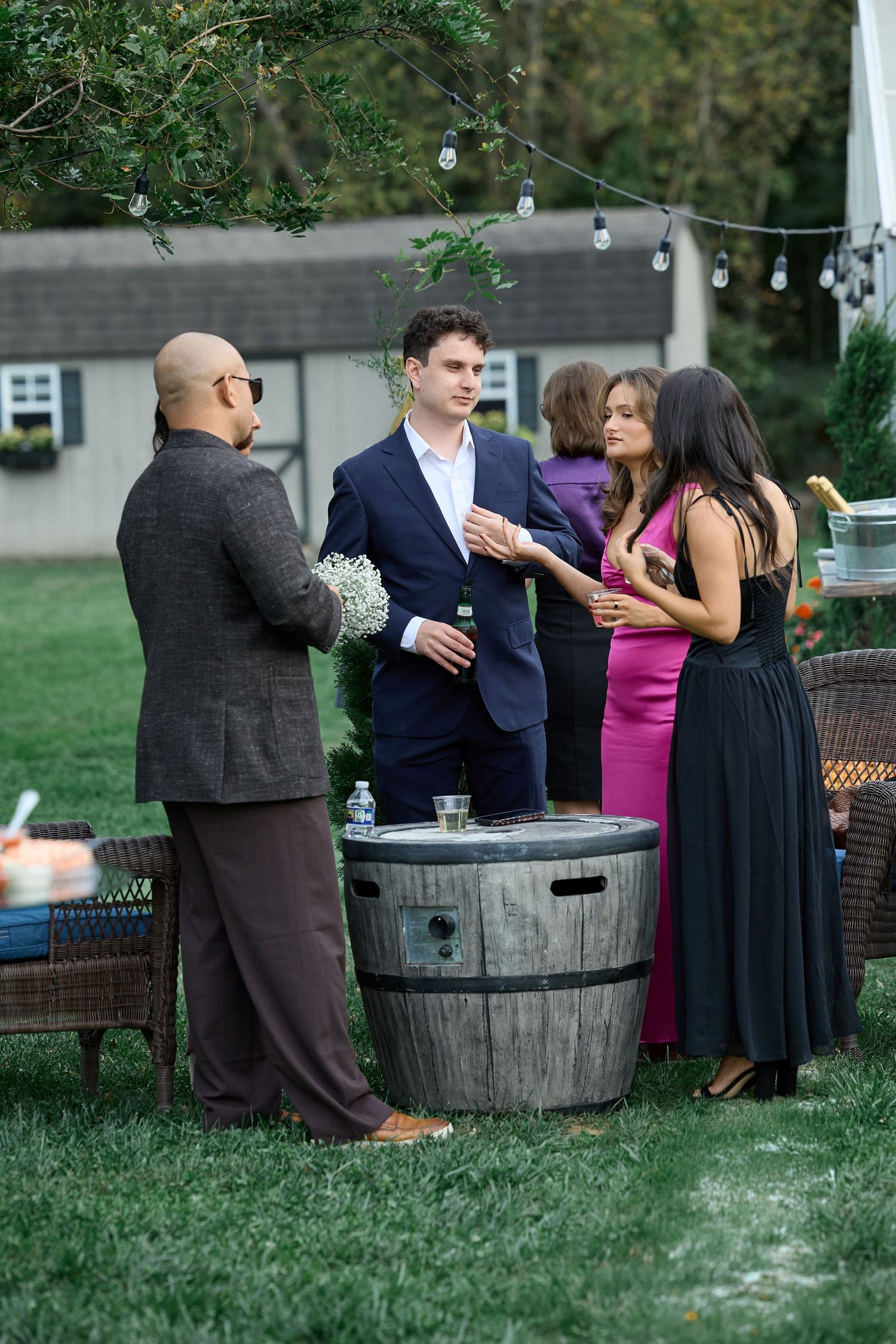 Guests in formal attire mingle around a wooden barrel table at an outdoor wedding reception under string lights.