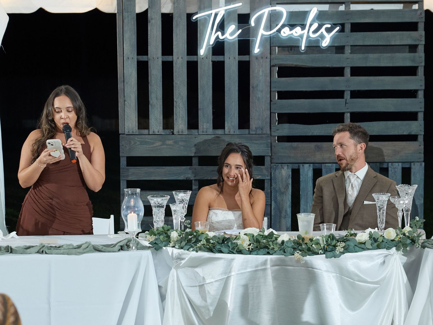 Bridesmaid gives a speech into a microphone while the emotional bride and groom listen at the head table beneath a neon The Pooles sign.