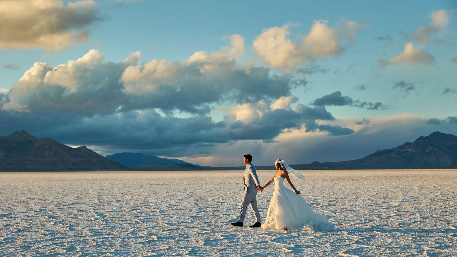 Tiny bride and groom stroll hand in hand across an expansive salt flat with towering clouds and mountains in the distance.