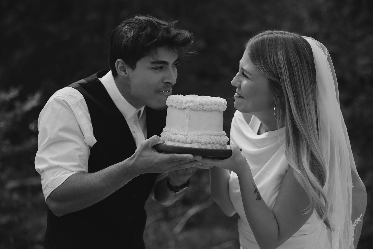 Black and white photo of a groom holding a small wedding cake while the veiled bride smiles at him.