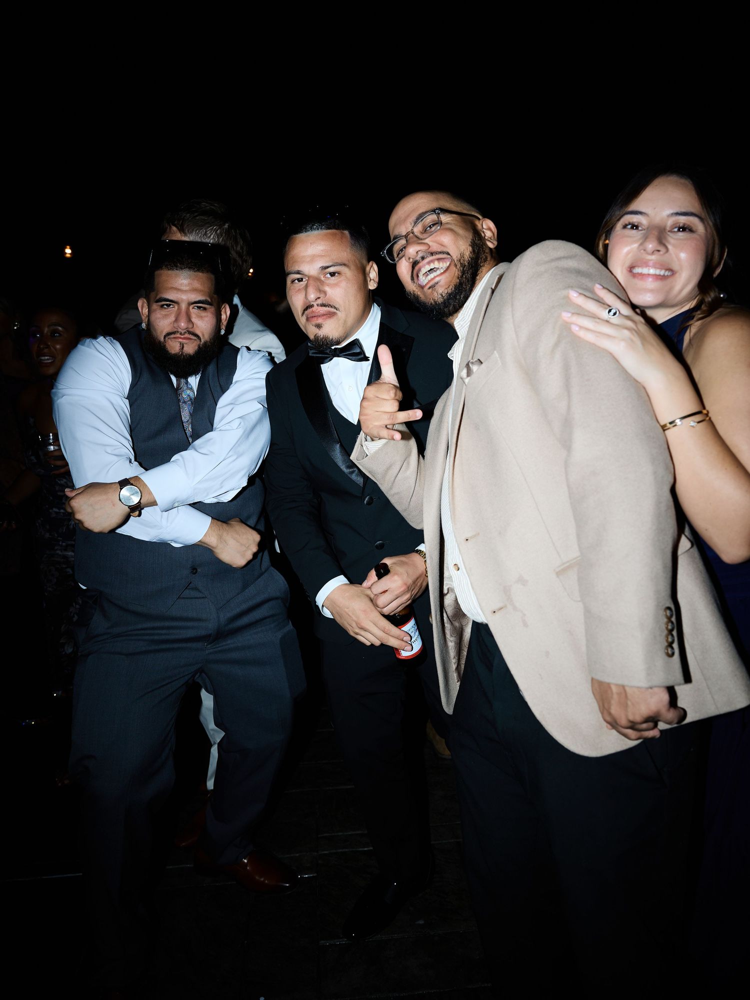 Four friends in dressy attire pose and smile together at a nighttime wedding celebration.