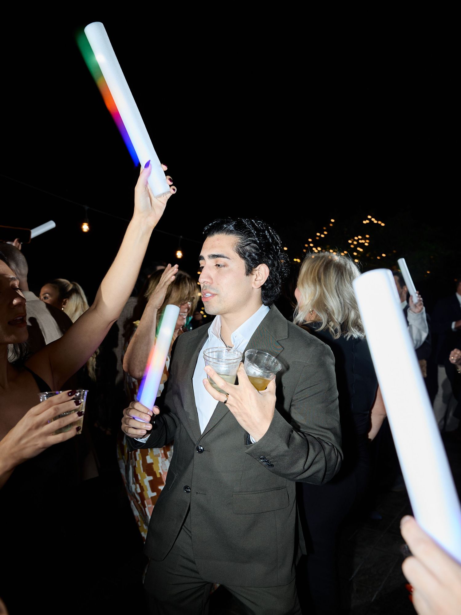 Man in a dark suit holds a cocktail while guests wave glowing foam sticks on the dance floor at a nighttime wedding.