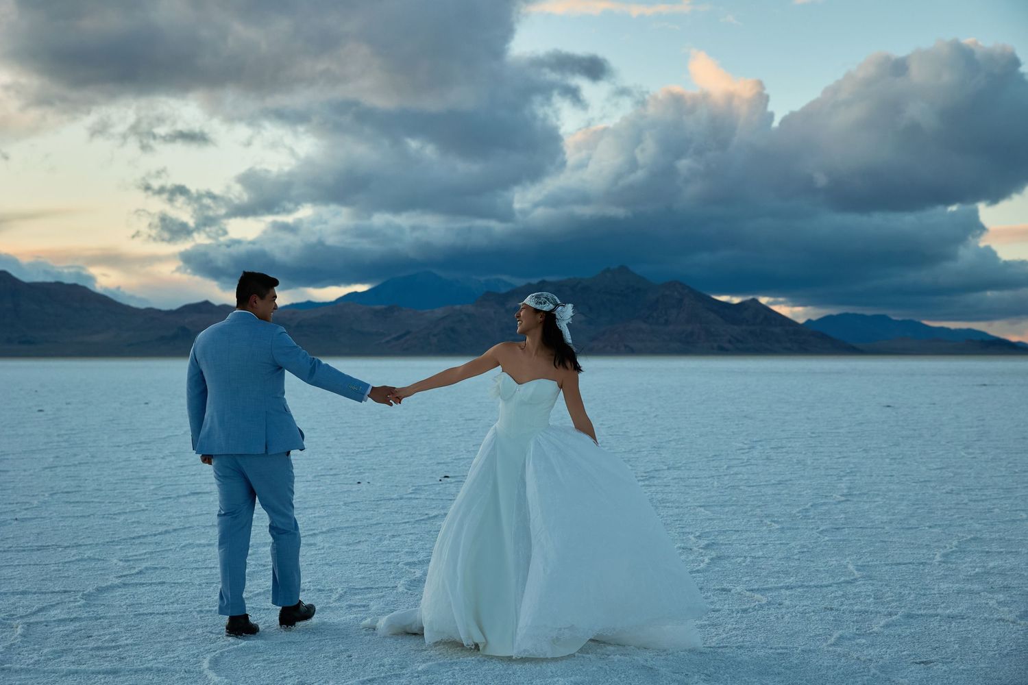 Groom in a blue suit leads the bride in a white gown by the hand across a reflective salt flat under a moody cloudy sky.
