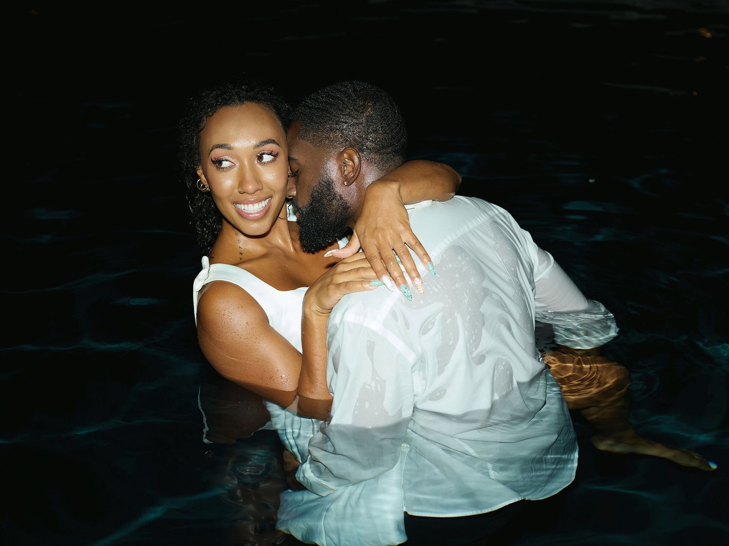 A smiling woman in a white dress hugs her bearded partner in a white shirt while standing in a dark nighttime pool.