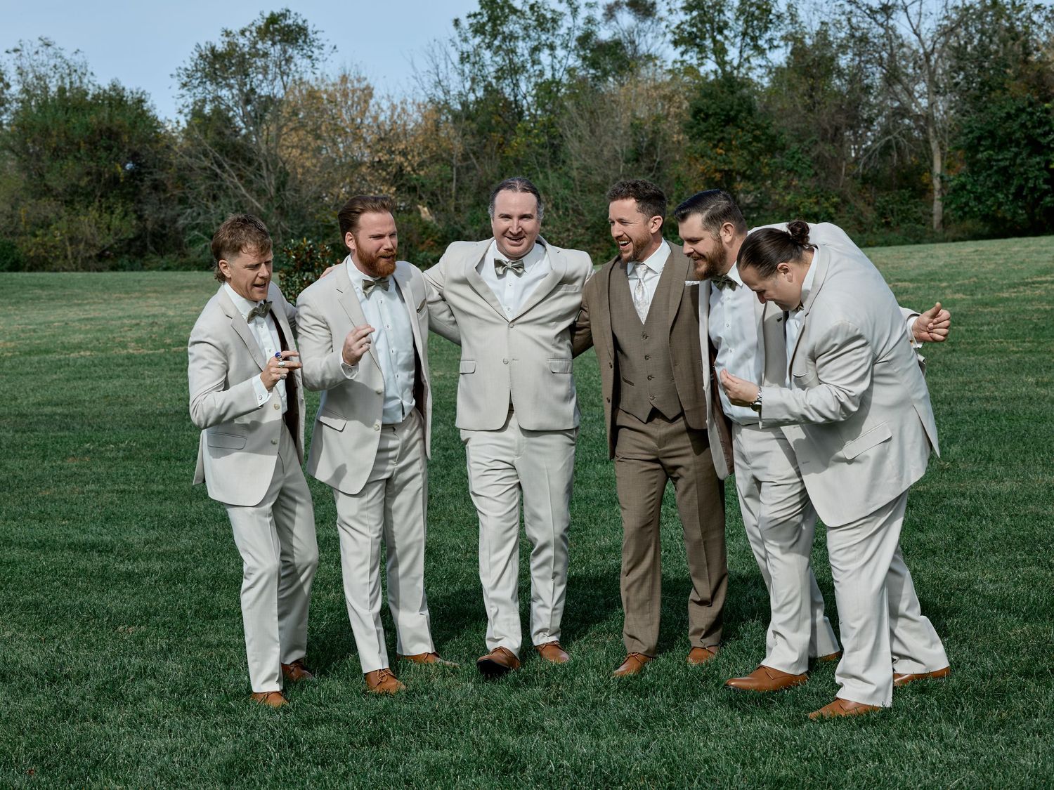 Groom in a brown vest laughs with four groomsmen in light gray suits while they link arms on a sunny grass lawn.