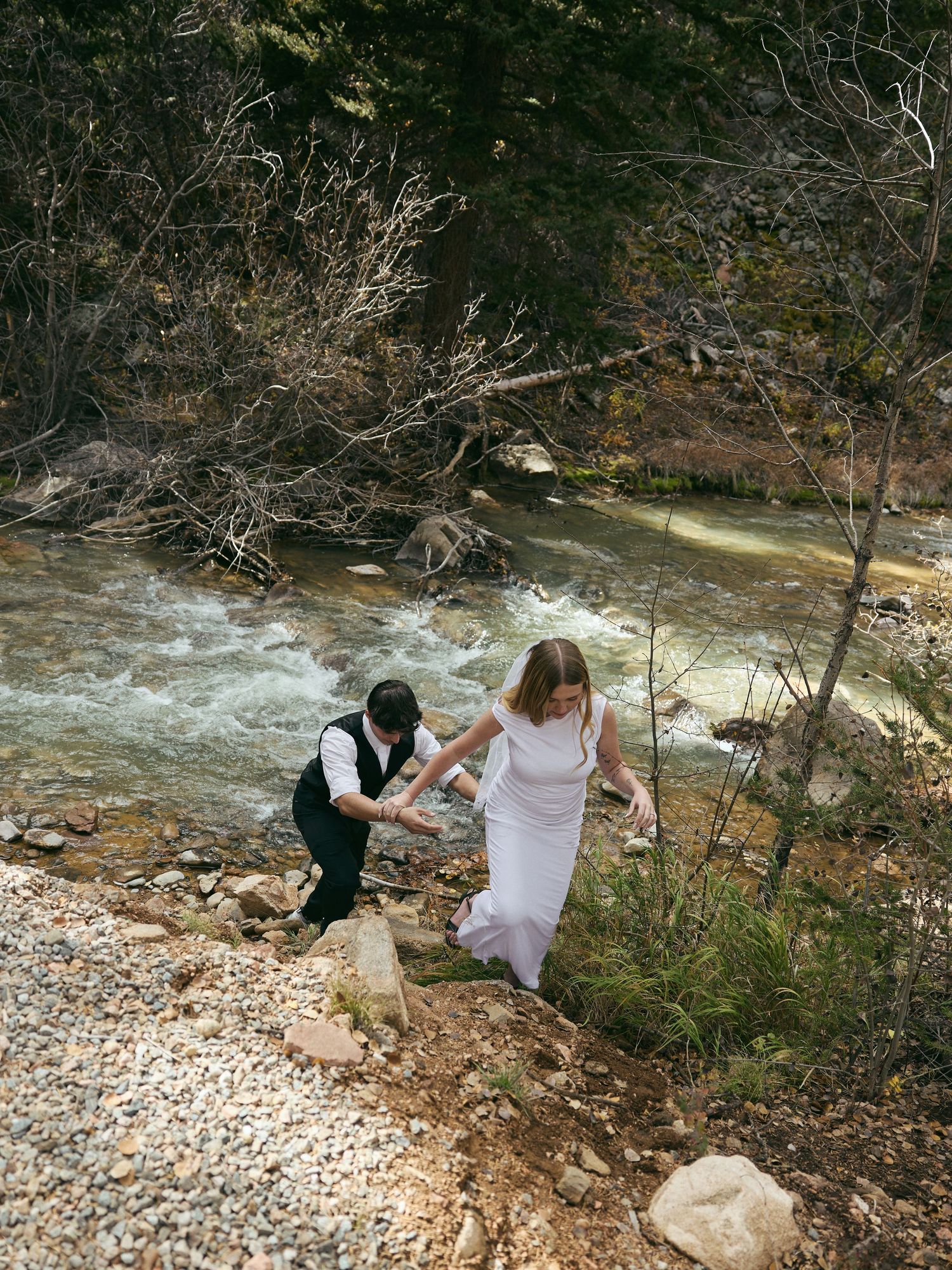 Groom helps his bride in a white dress climb up a rocky riverbank beside a rushing mountain stream during their elopement.