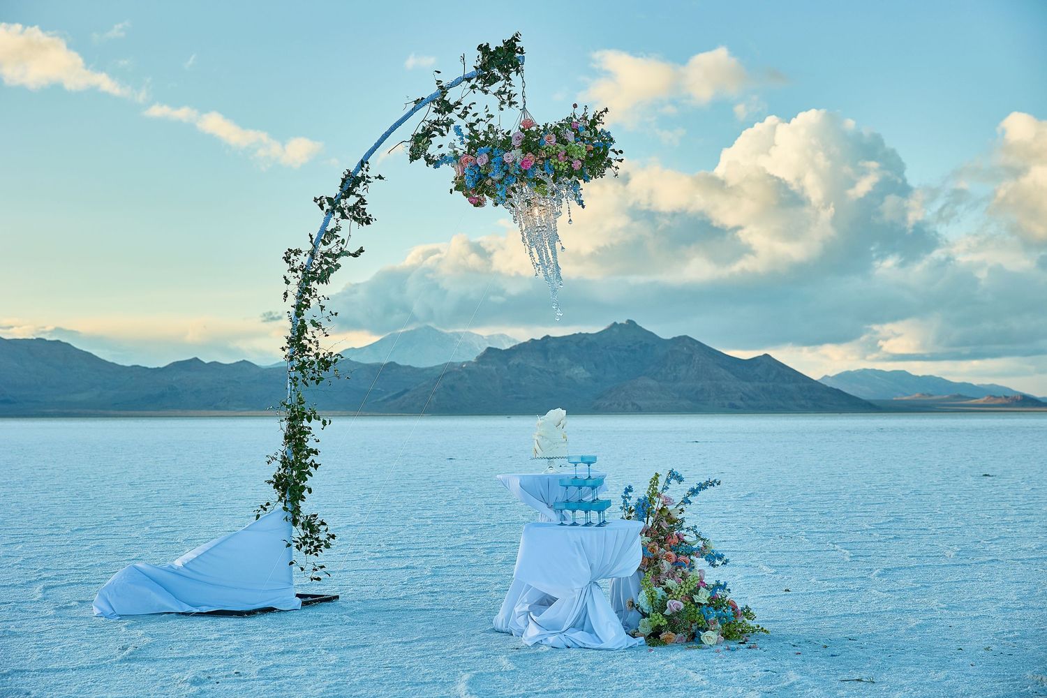 Floral wedding arch with greenery and a crystal chandelier stands beside a cake table on a vast white salt flat at sunset.