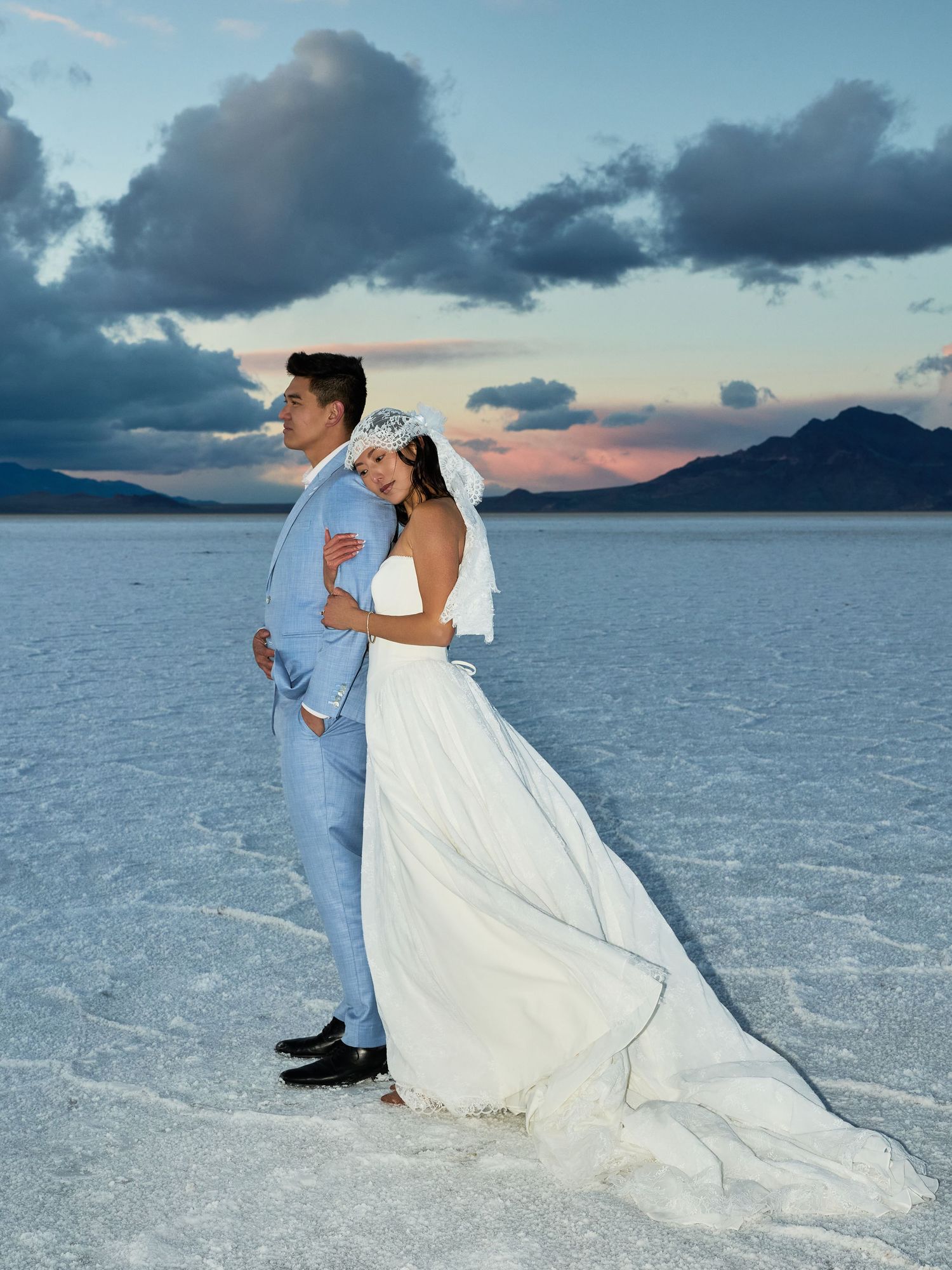Bride in a long white gown and groom in a blue suit stand back to back on a cracked salt flat beneath moody clouds.