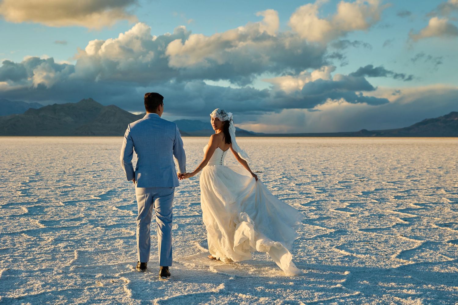 Bride in a flowing white gown and groom in a light blue suit hold hands and walk across a vast white salt flat under dramatic clouds.