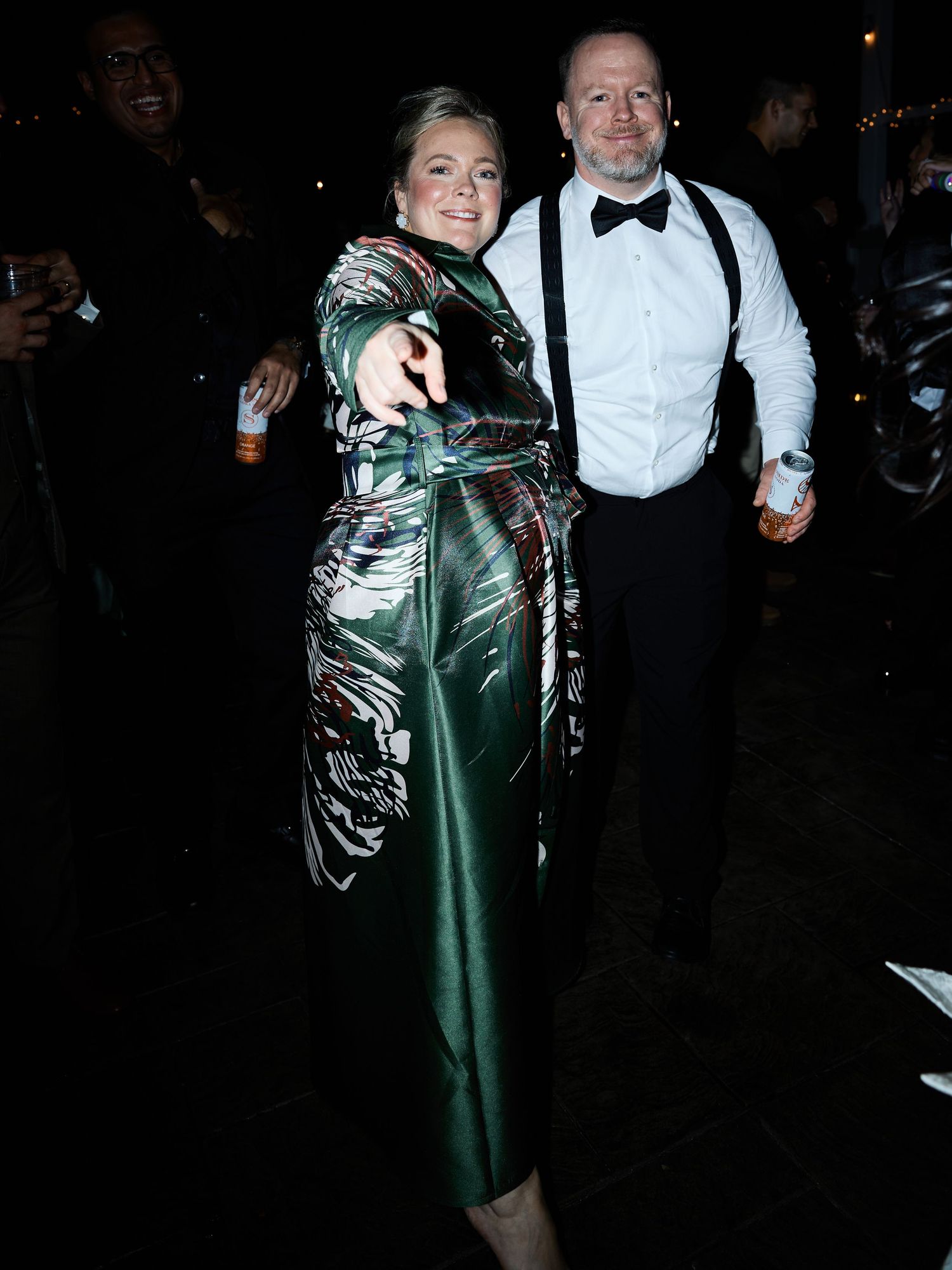 A woman in a green patterned satin dress points at the camera beside a man in suspenders and a bow tie at a party.