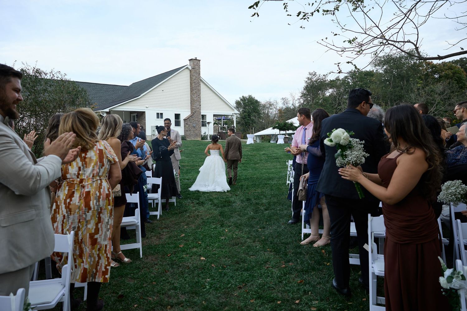 The bride walks down a grassy aisle toward a country house as guests holding bouquets stand and applaud on both sides.