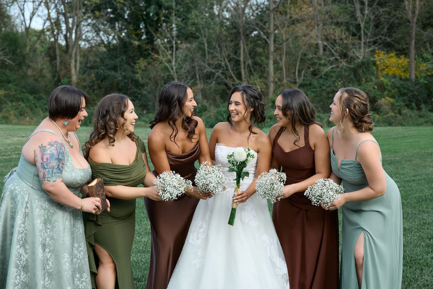 Smiling bride in a white gown stands arm in arm with four bridesmaids in sage and brown dresses holding white bouquets outdoors.