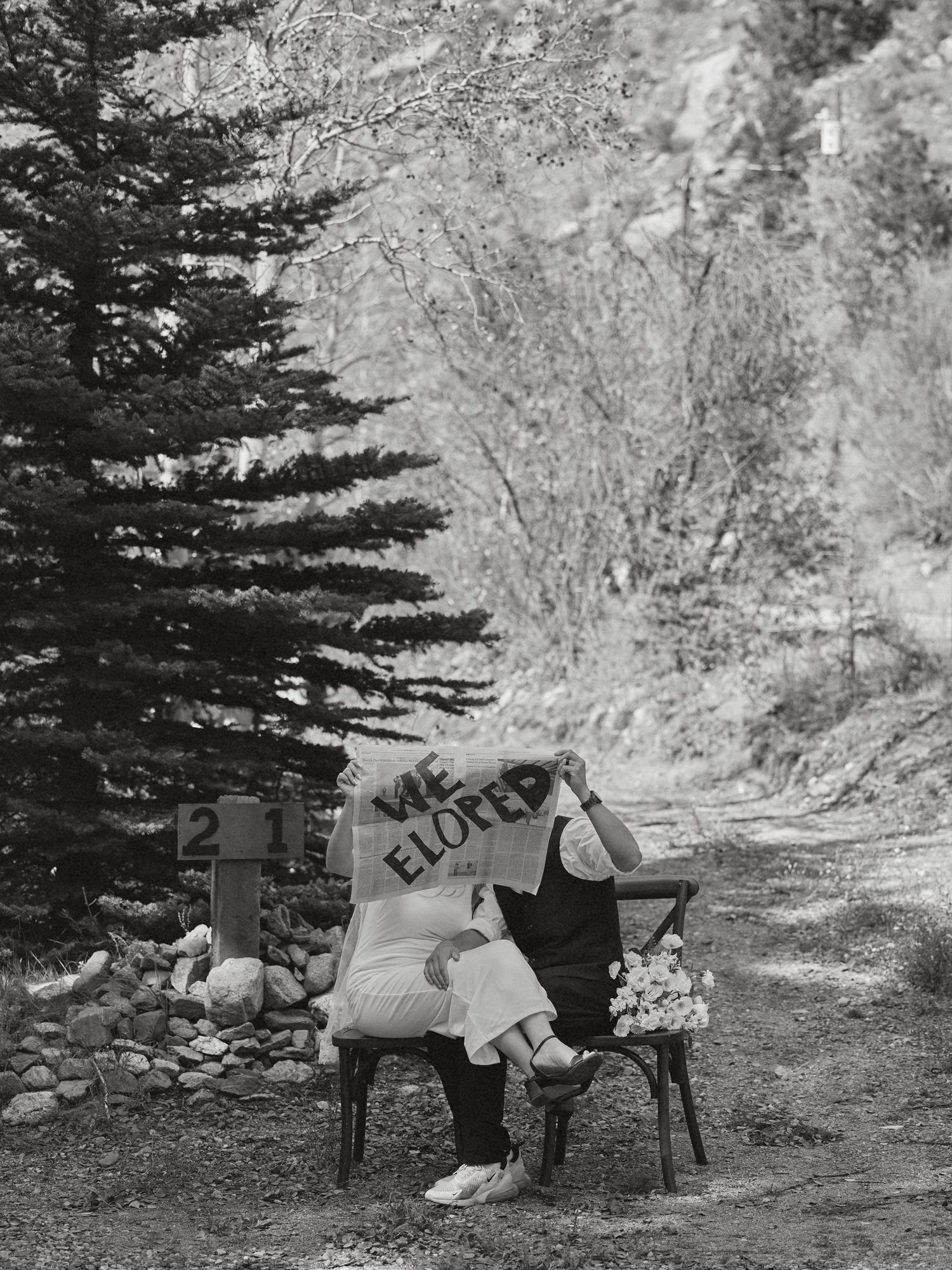 Black and white photo of an elopement couple seated on chairs hiding behind a We Eloped sign beside a pine tree and bouquet.