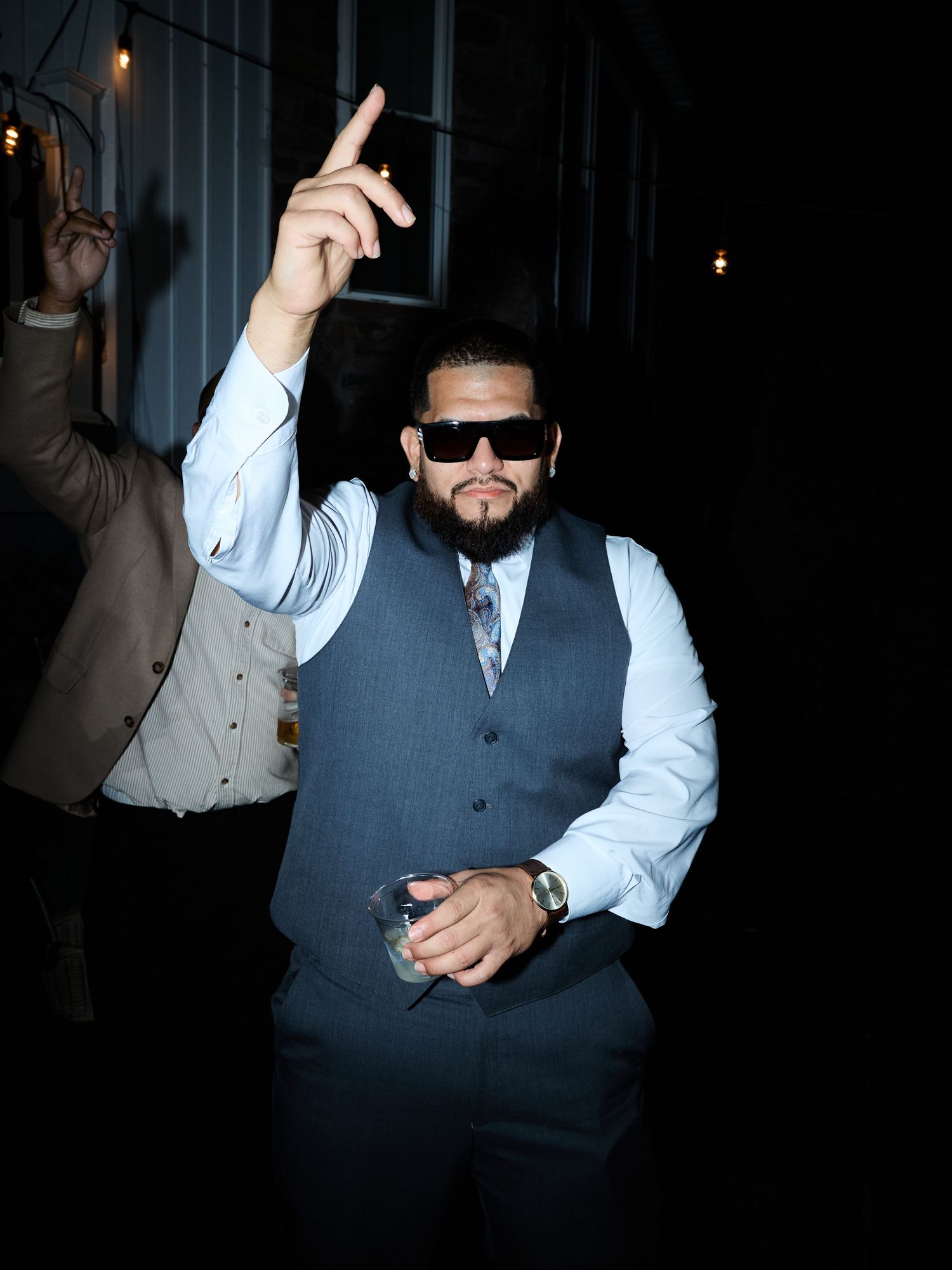A bearded man in sunglasses and a gray vest points upward while dancing and holding a drink at an evening party.