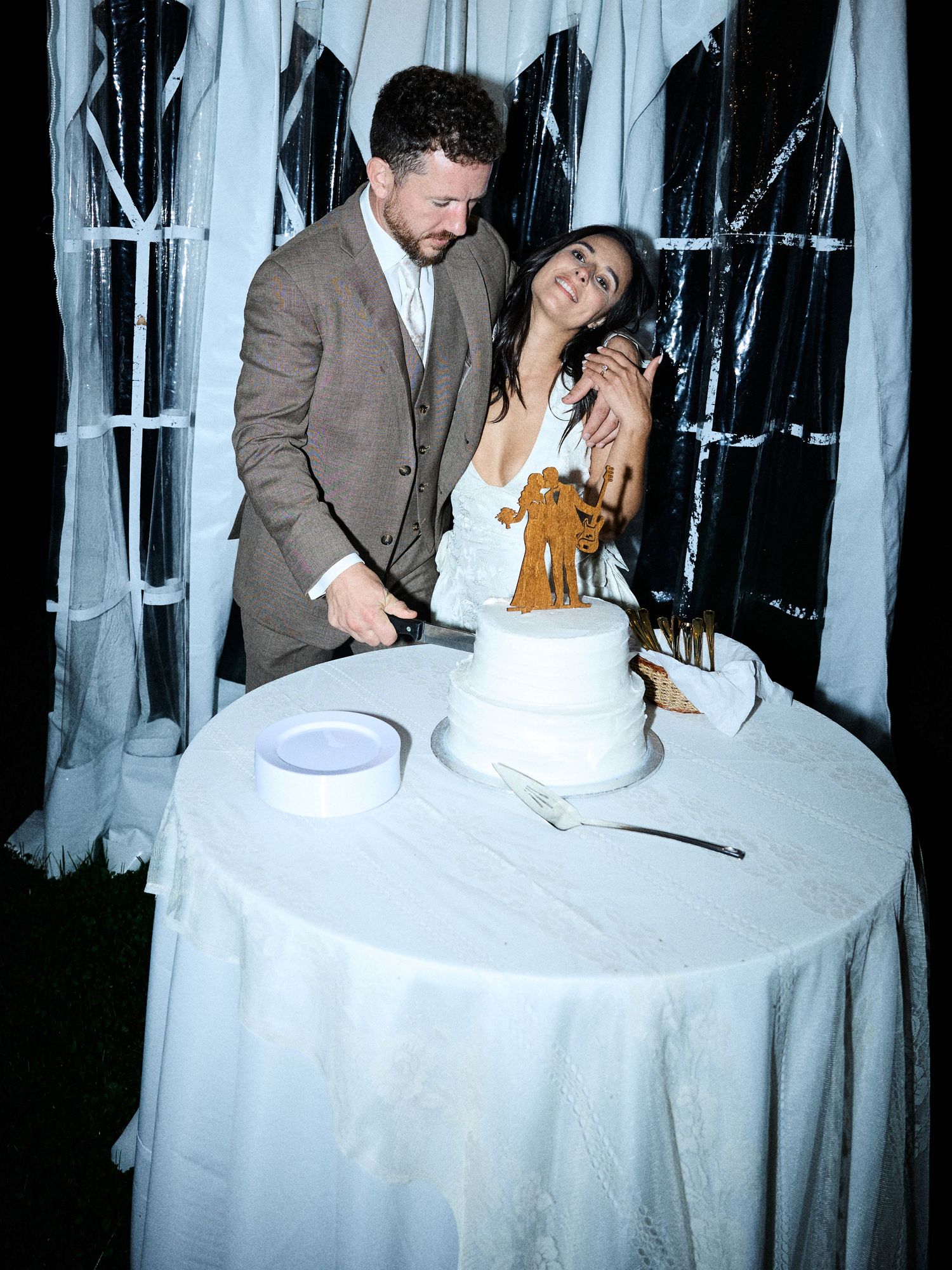 A smiling bride and groom pose together behind their white tiered wedding cake with a wooden topper during a nighttime reception.