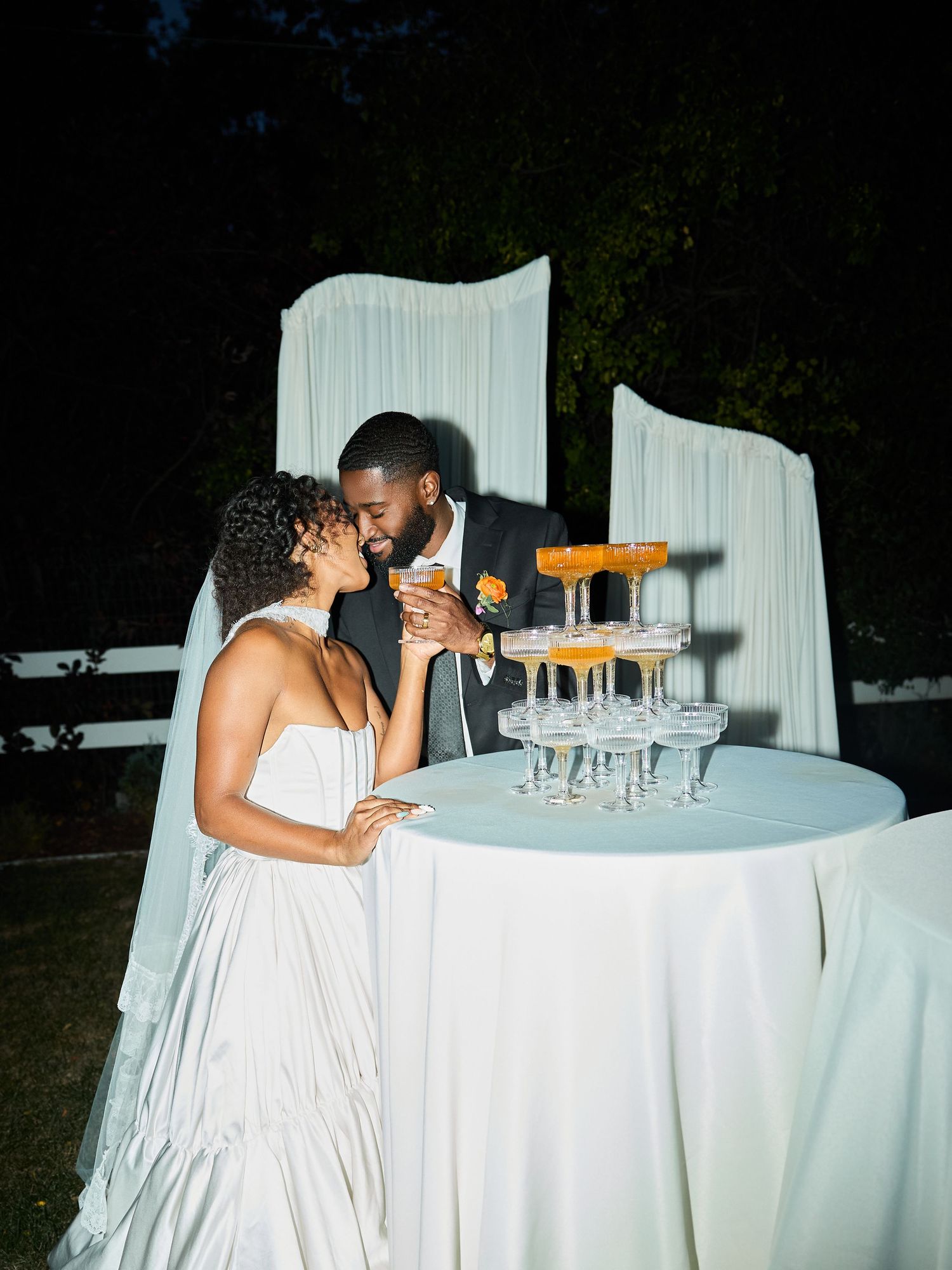 A groom in a dark suit nuzzles his bride in a flowing white gown beside a champagne tower at their nighttime outdoor wedding reception.