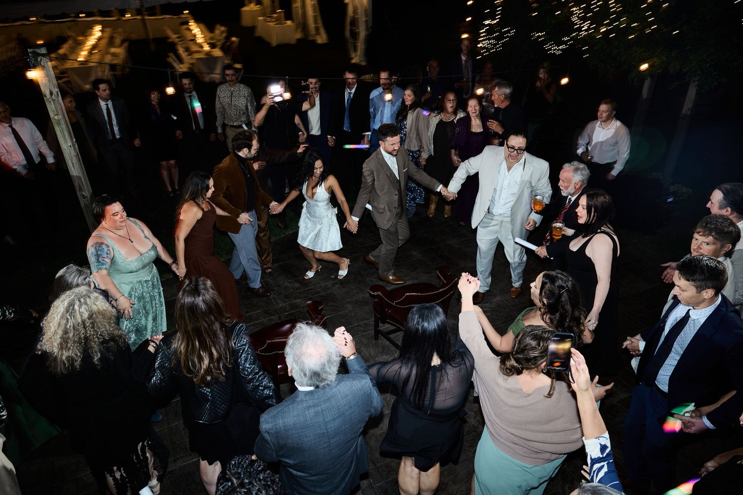 Wedding guests dance and cheer on an outdoor patio at night under warm string lights during the reception.