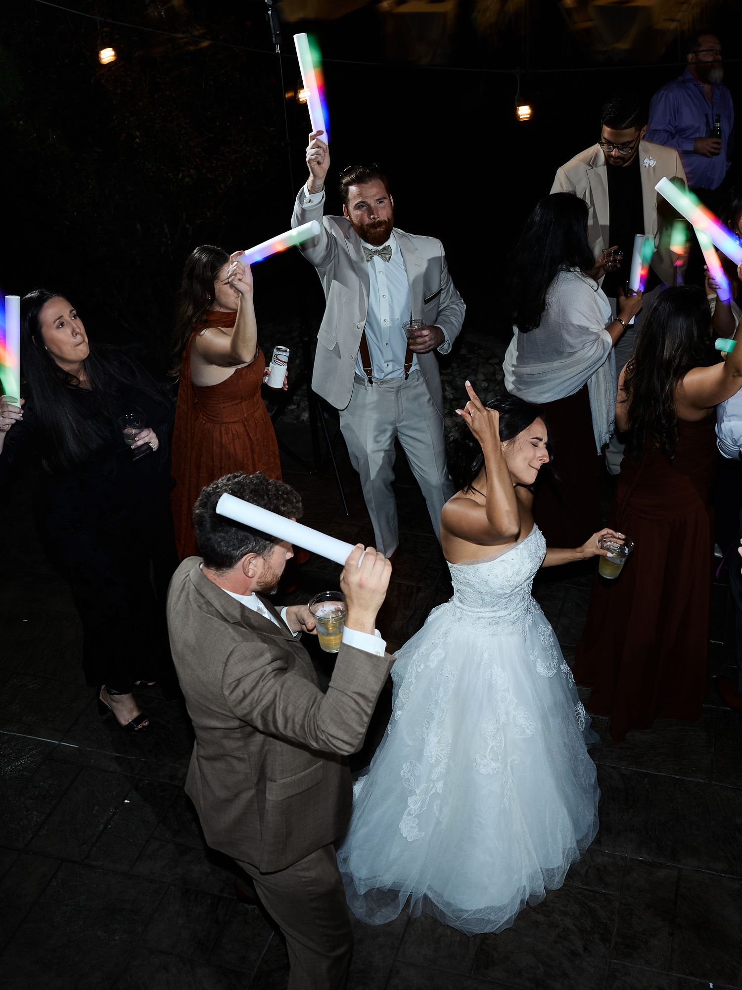 Wedding guests and a bride in a white tulle dress dance under glowing light sticks on an outdoor dance floor at night.