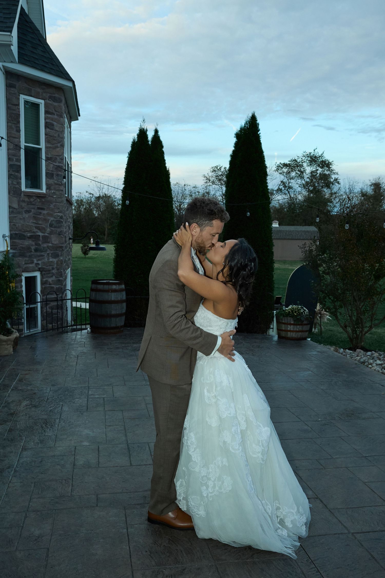 A bride and groom share a romantic kiss on a stone patio at dusk beside a stone house and tall evergreen trees.