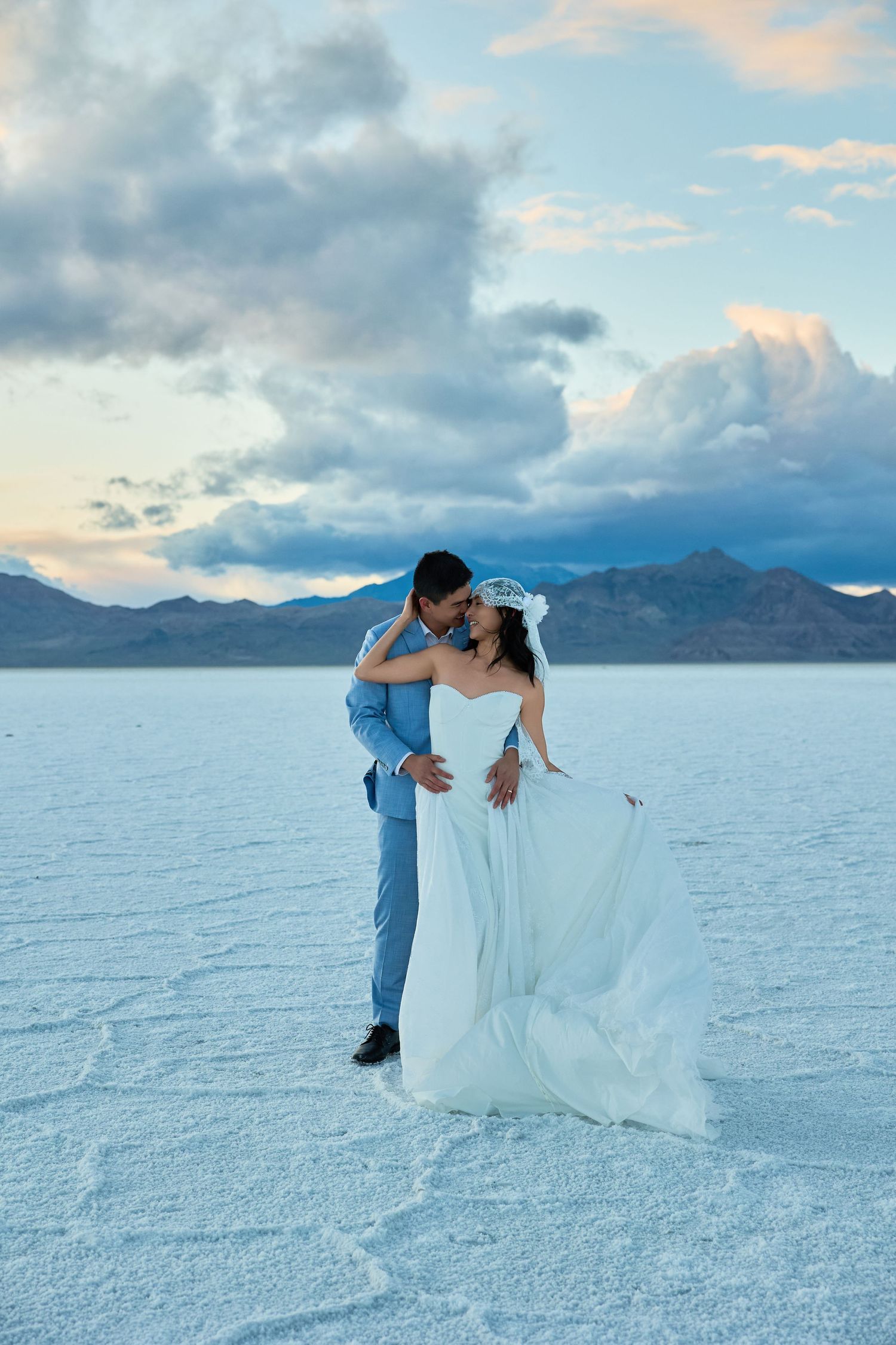 Couple leans in for a kiss on a white salt flat, the bride in a flowing white gown and the groom in a blue suit.