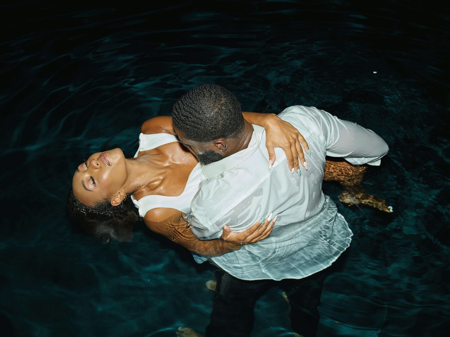 Overhead view of a man in a white shirt dipping a woman backward in dark pool water during a romantic night photoshoot.