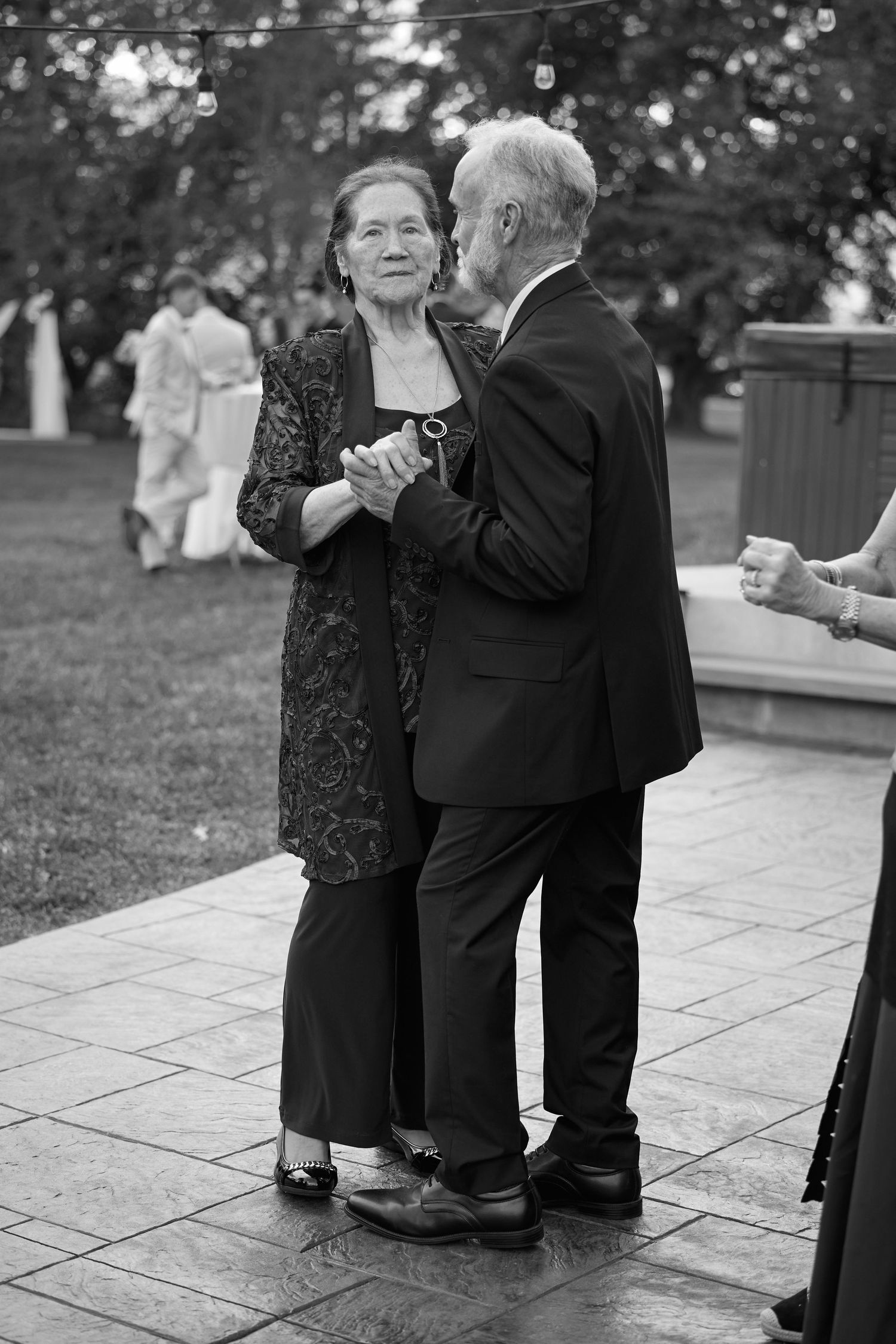 Black and white photo of an elderly couple in formal wear slow dancing together on a stone patio at an outdoor celebration.