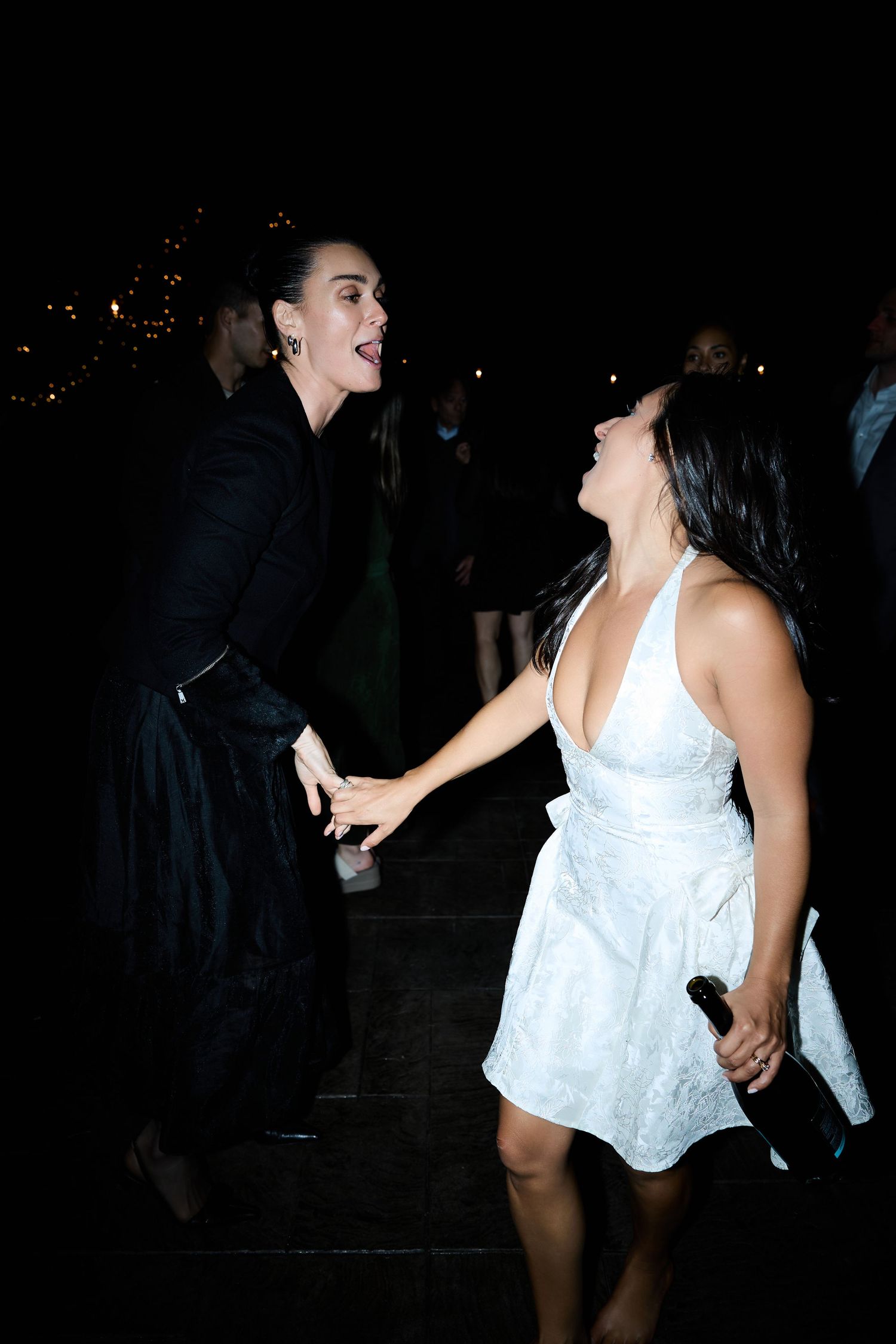A woman in a white dress holding a bottle dances and laughs with a woman in black at a nighttime reception.