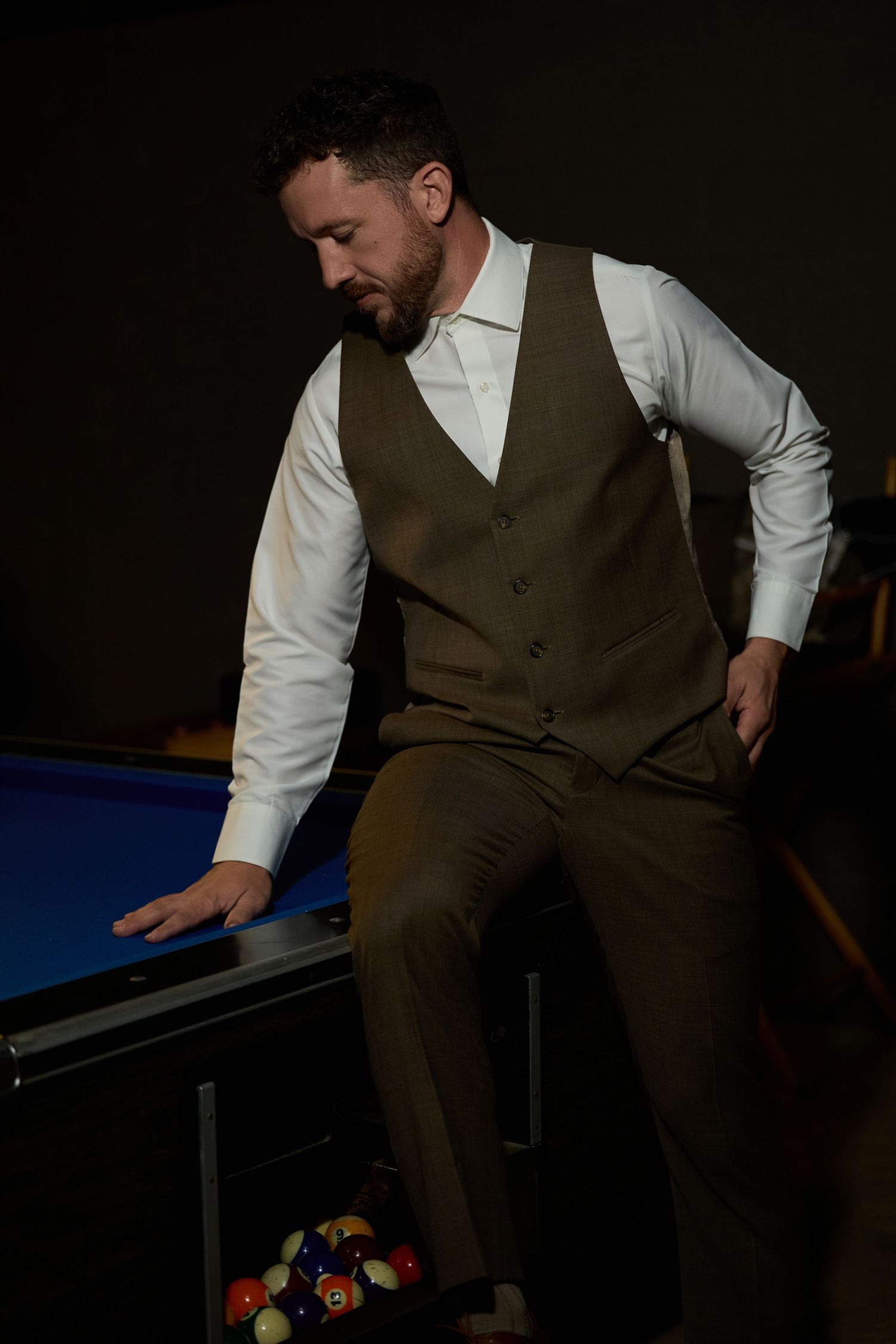 Bearded groom in a brown tweed vest and white shirt leans against a pool table in a dimly lit billiards room.
