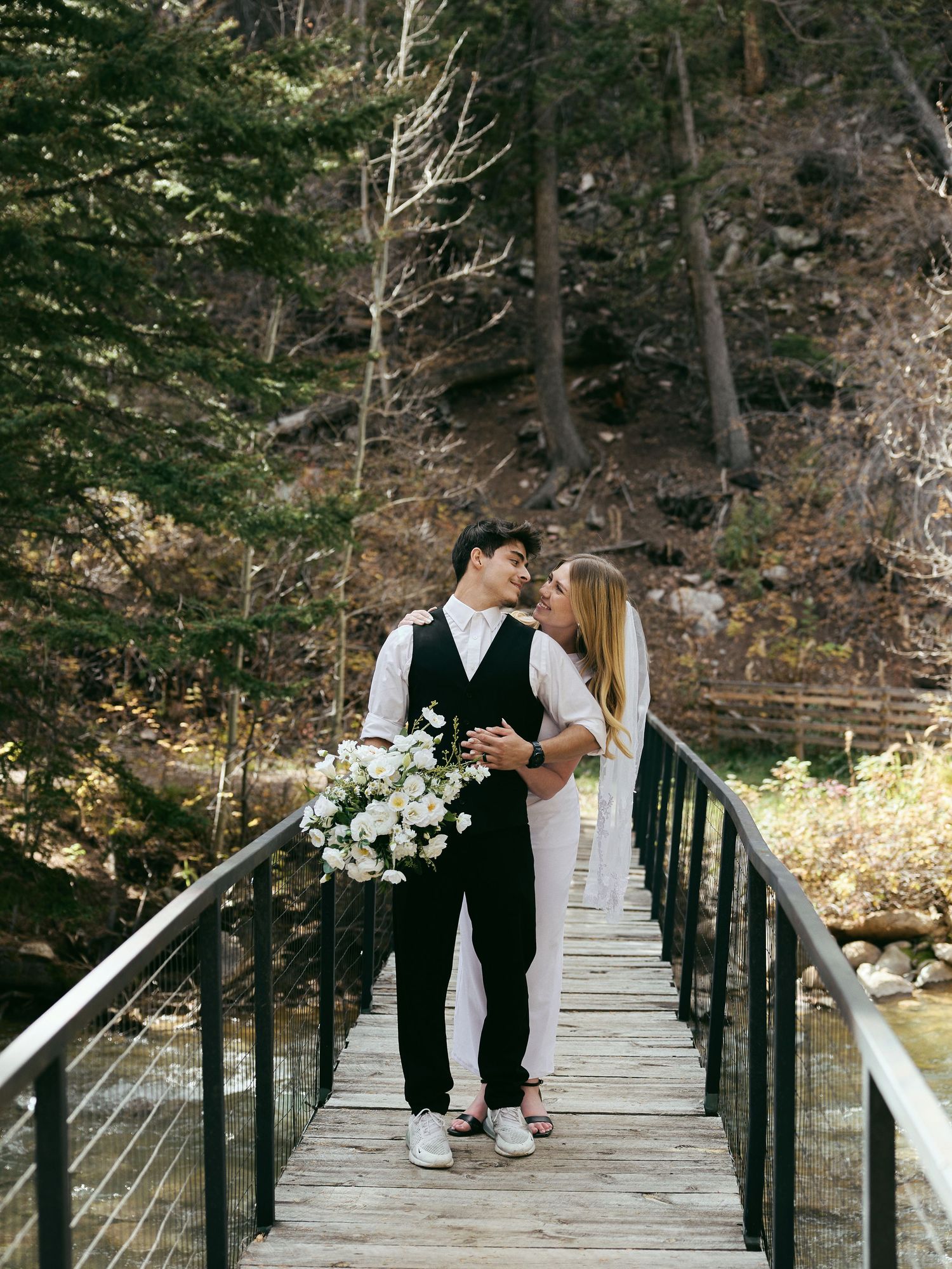 Smiling groom embraces his bride holding a white floral bouquet on a wooden bridge in a mountain forest elopement.