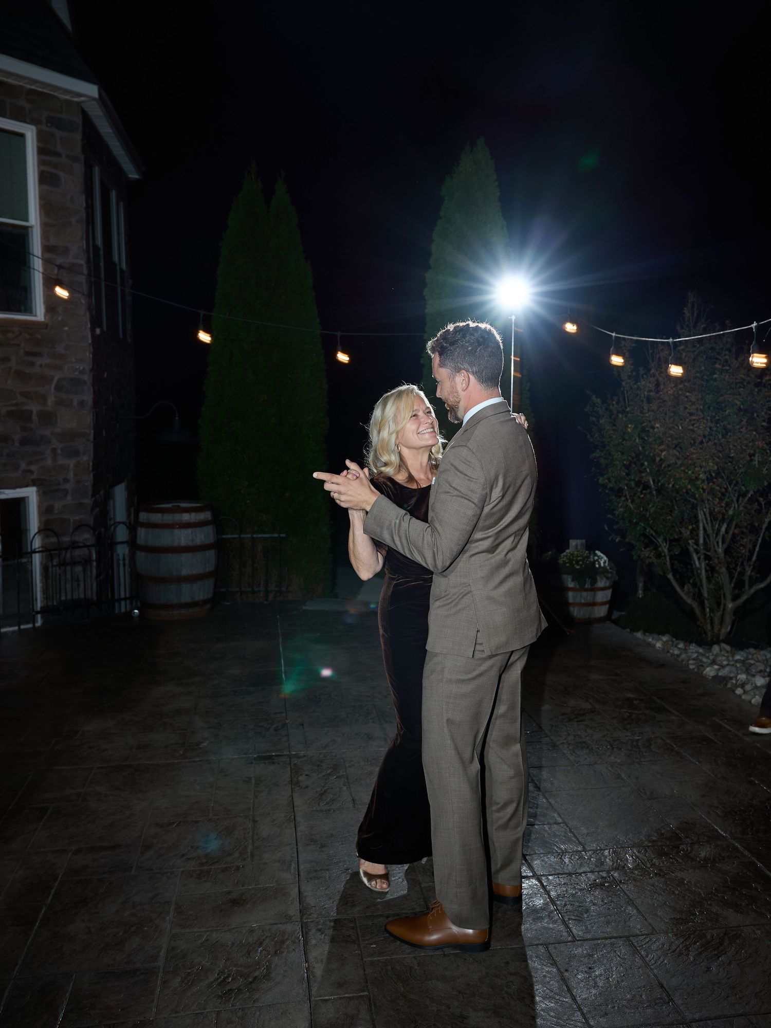 A groom in a tan suit dances with a blonde woman in a black dress under string lights on a patio at night.