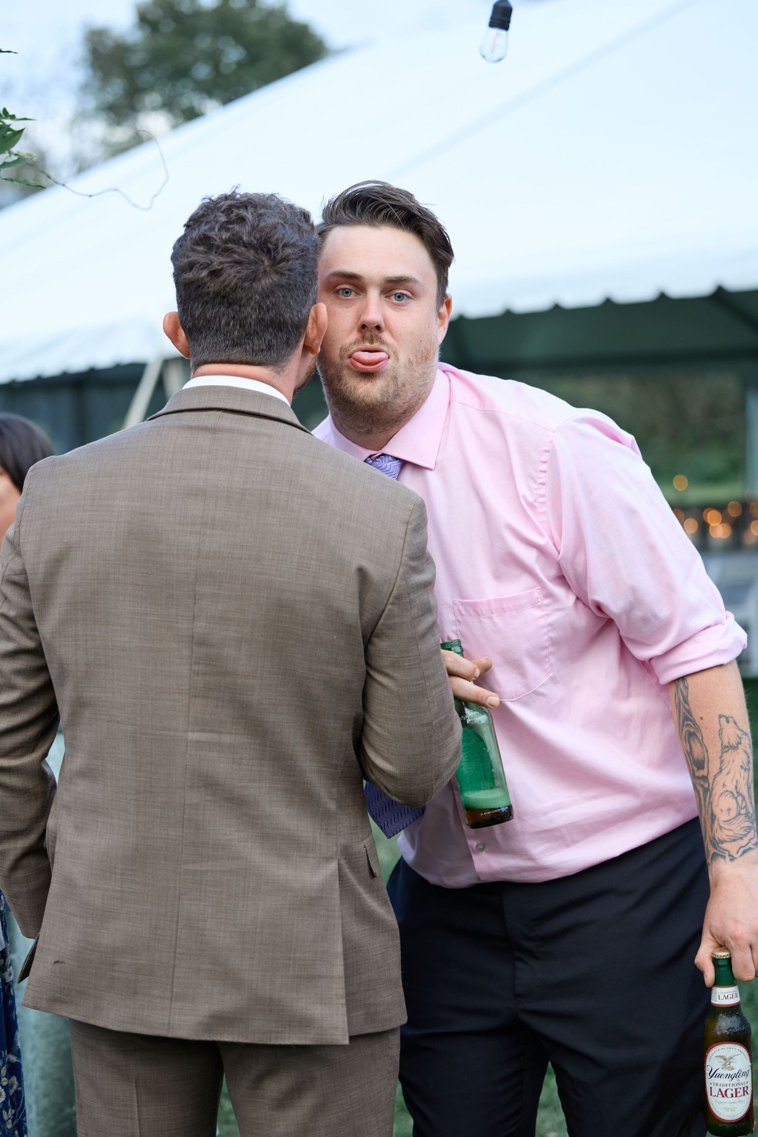 Two men in a pink dress shirt and tweed suit share a playful moment holding beer bottles at an outdoor tented wedding reception.