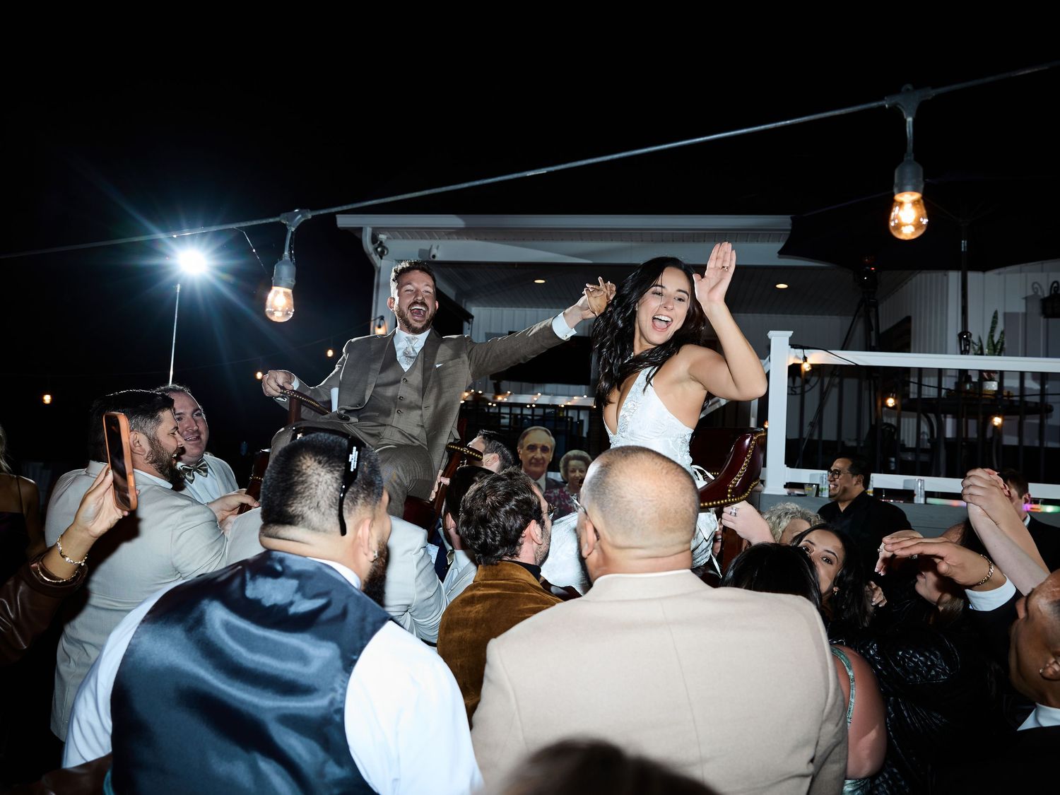 Wedding guests raise the smiling bride and groom on chairs in a joyful hora under string lights at night.