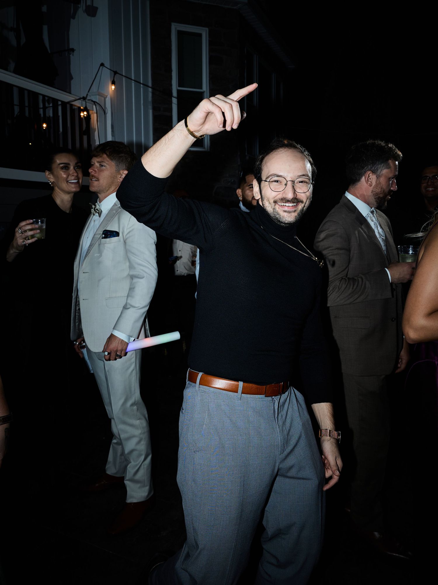 A smiling man in glasses and a black sweater points upward while dancing at an outdoor evening wedding celebration.